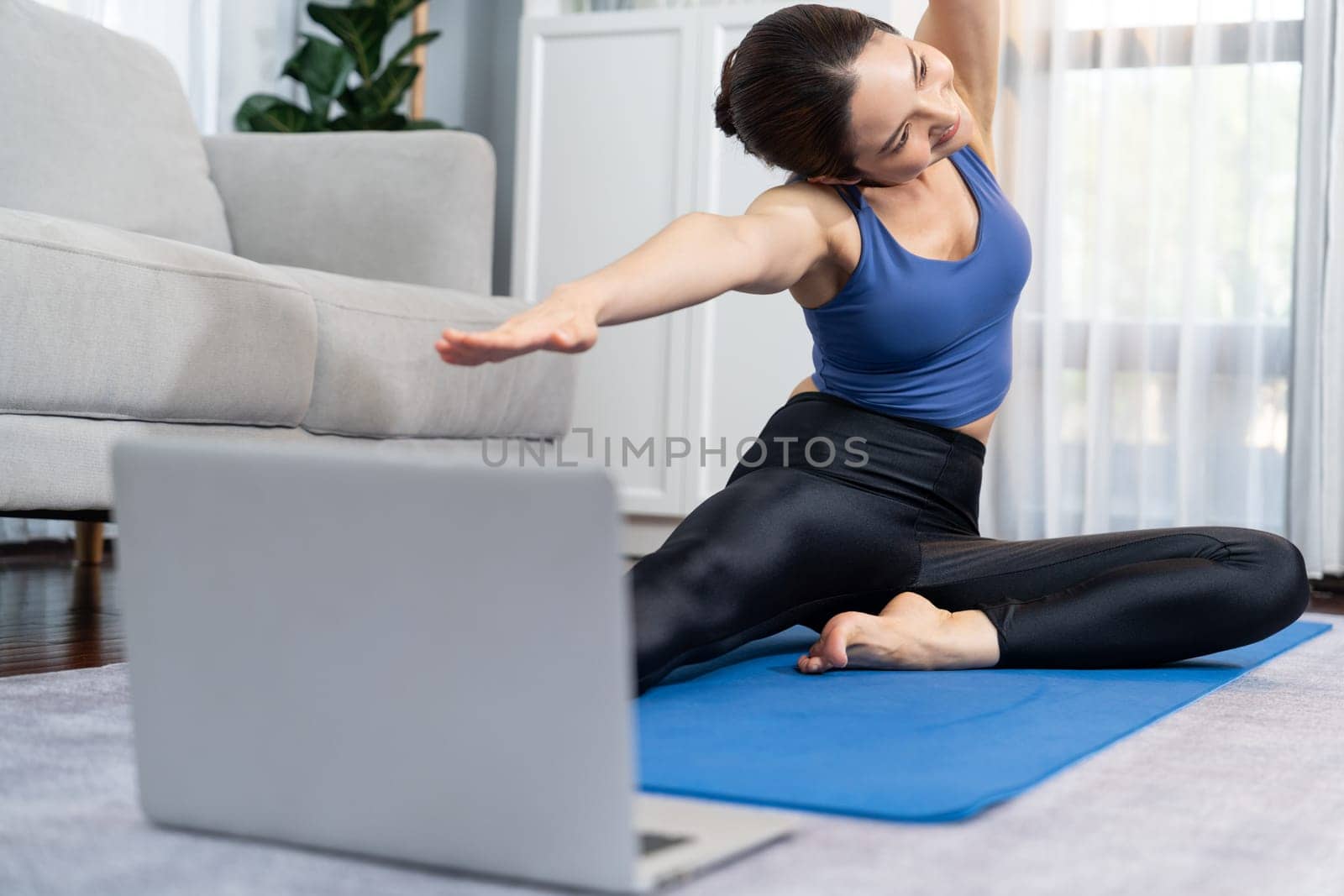 Focused laptop on the floor showing online exercise training video, while sporty athletic woman concentrate on warm-up and stretching routine in blurred background. Vigorous