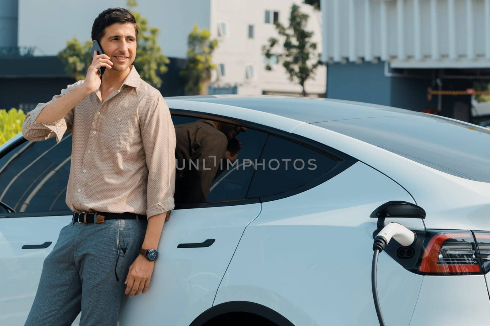 Young man recharge EV electric vehicle at green city commercial mall parking lot while talking on phone. Sustainable urban lifestyle for eco friendly EV car with battery charging station. Expedient