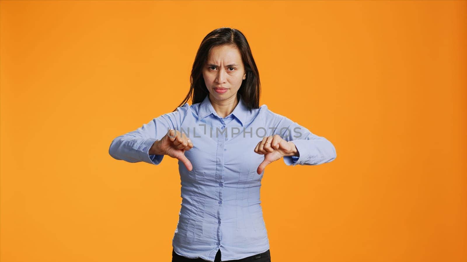 Asian woman giving dislike gesture on camera, showing disagreement and negativity over orange background. Negative unhappy adult presenting thumbs down symbol in studio, wrong idea.