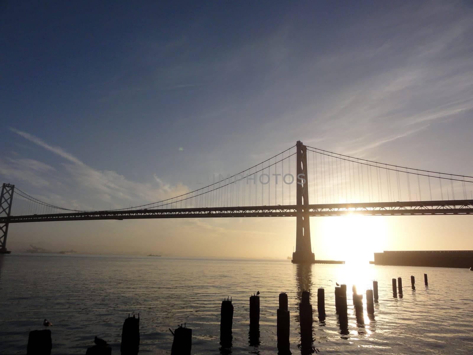 A stunning photo of a sunrise through the Bay Bridge, a suspension bridge that connects San Francisco and Oakland in California. The photo shows the bridge silhouetted against the golden orange sunrise, creating a contrast between the dark and the light. The sunrise is shining through the bridge, creating a geometric pattern of light and shadow. The sky is a light blue color with some clouds. The water is a dark blue color and is calm. There are some wooden pilings in the foreground, adding a touch of rustic charm to the scene. The photo captures the beauty and the elegance of the Bay Bridge