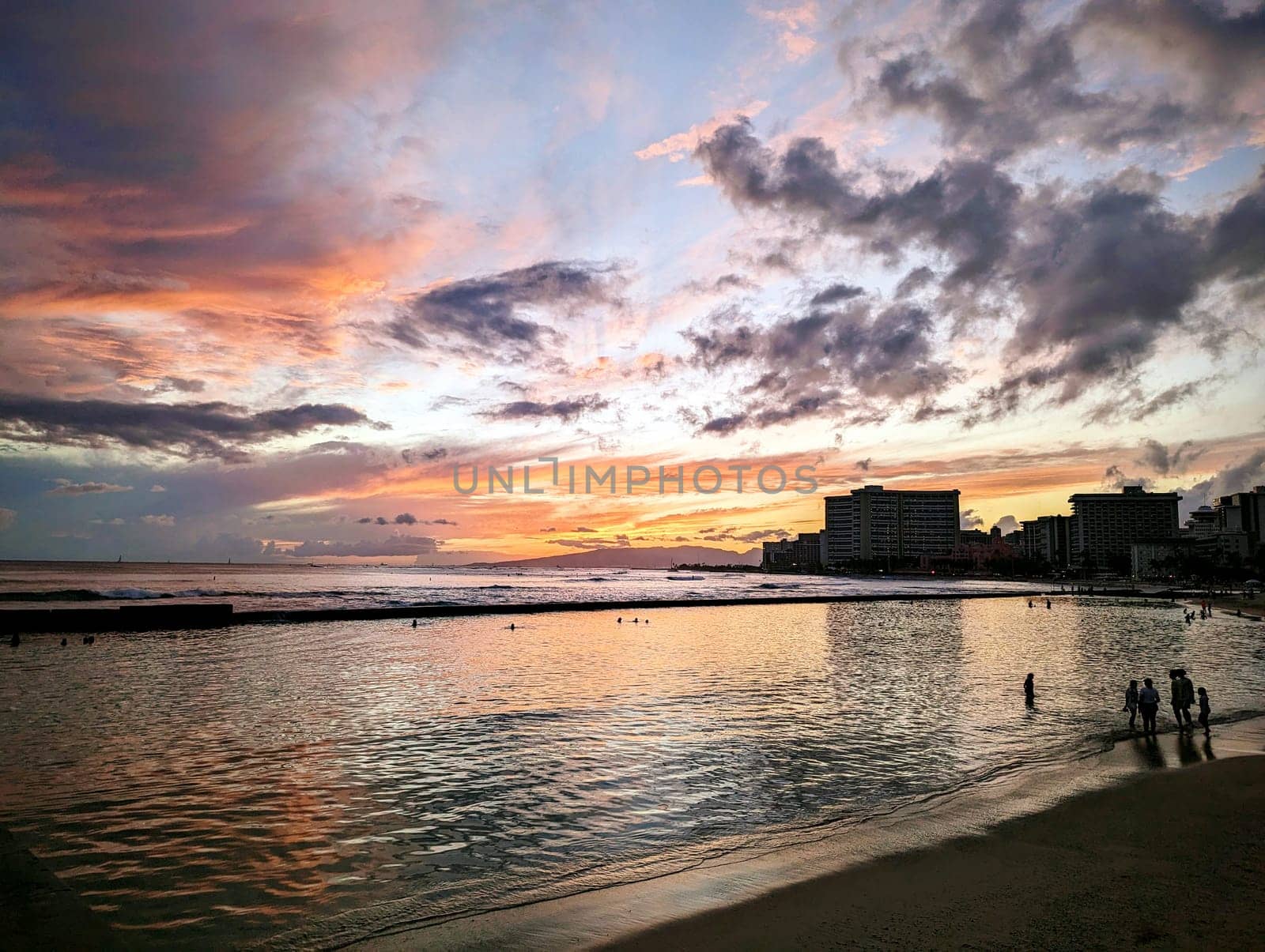 Waikiki - May 29, 2023: A stunning view of the sun setting over the Pacific Ocean, as seen from the sandy shore of Waikiki Beach in Honolulu, Hawaii. The sky is painted with vibrant colors and the water reflects the light. The silhouette of the city skyline and the palm trees add contrast and interest to the scene