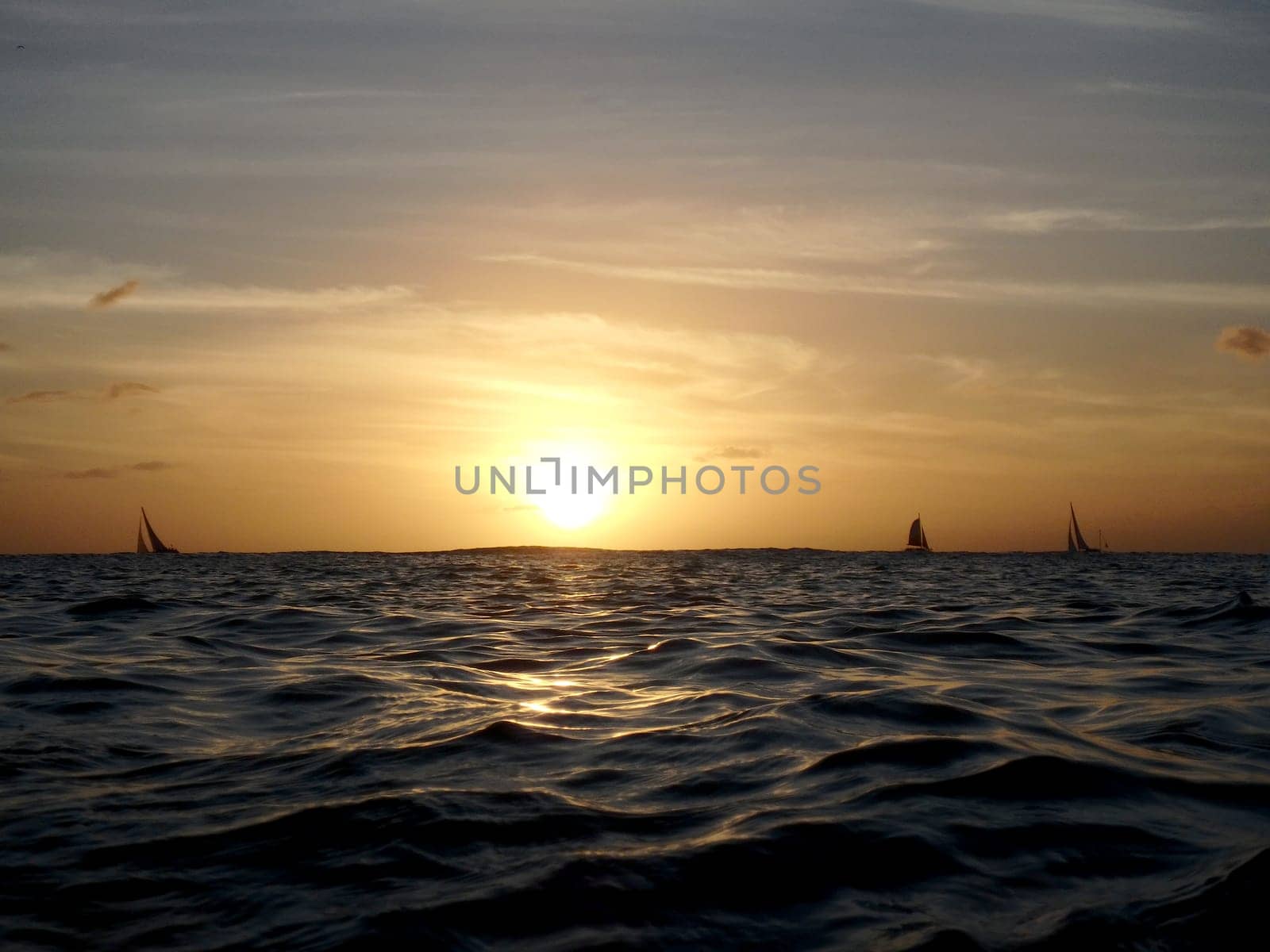 Stunning sunset over the ocean in Waikiki, Hawaii. The sun is setting over the horizon and is partially obscured by clouds. The sky is orange and yellow with a few clouds. The ocean is dark and choppy with a few sailboats in the distance.