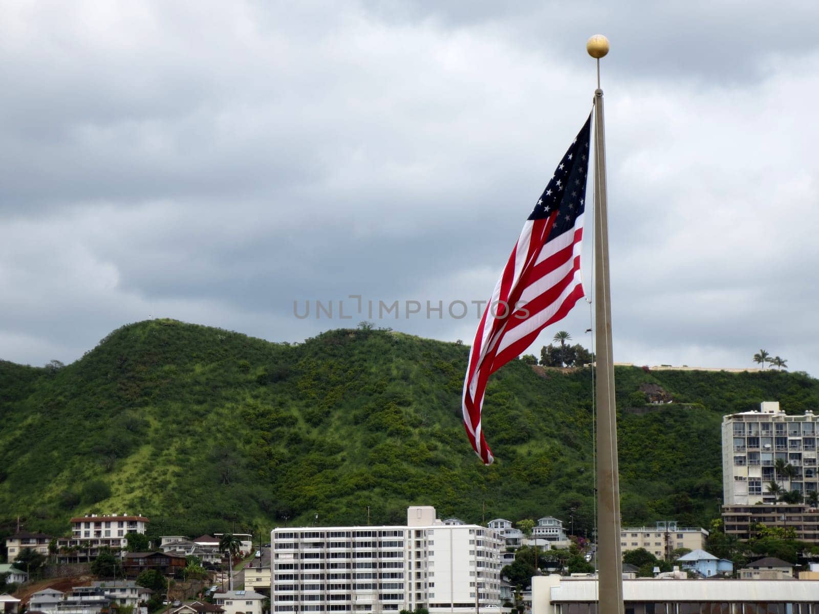 Honolulu - March 15, 2013: American flag flying on a flagpole in front of Punchbowl, a volcanic crater and a national memorial cemetery in Honolulu, Hawaii. The flag is at full mast and is waving in the wind. The background consists of a mountainous landscape with buildings and trees. The sky is overcast.