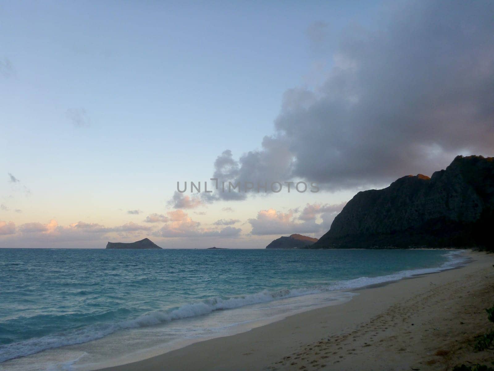 A tranquil photo of Waimanalo Beach, a scenic beach on the east coast of Oahu, Hawaii. The photo shows the ocean and the beach at dusk, with a soft light and a gentle breeze. The ocean is a blue-green color with white waves crashing onto the shore. The beach is sandy and appears to be empty. The sky is a light blue with a few clouds. The background consists of a mountain range with a few palm trees. The photo captures the beauty and the calmness of nature.