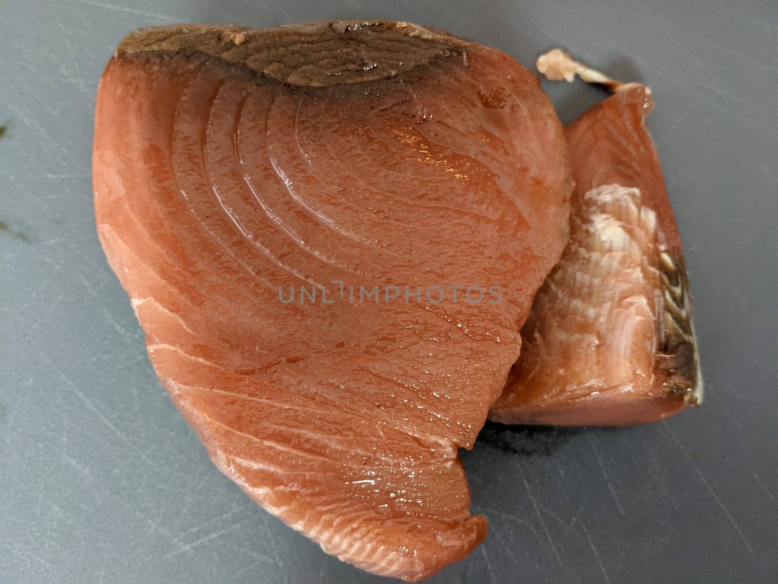 Two fresh fish fillets on a gray surface, ready to be cooked. The fillets are pink in color and have a shiny texture. The fillets are cut in a way that they are ready to be cooked. The background is a gray surface, possibly a kitchen countertop.
