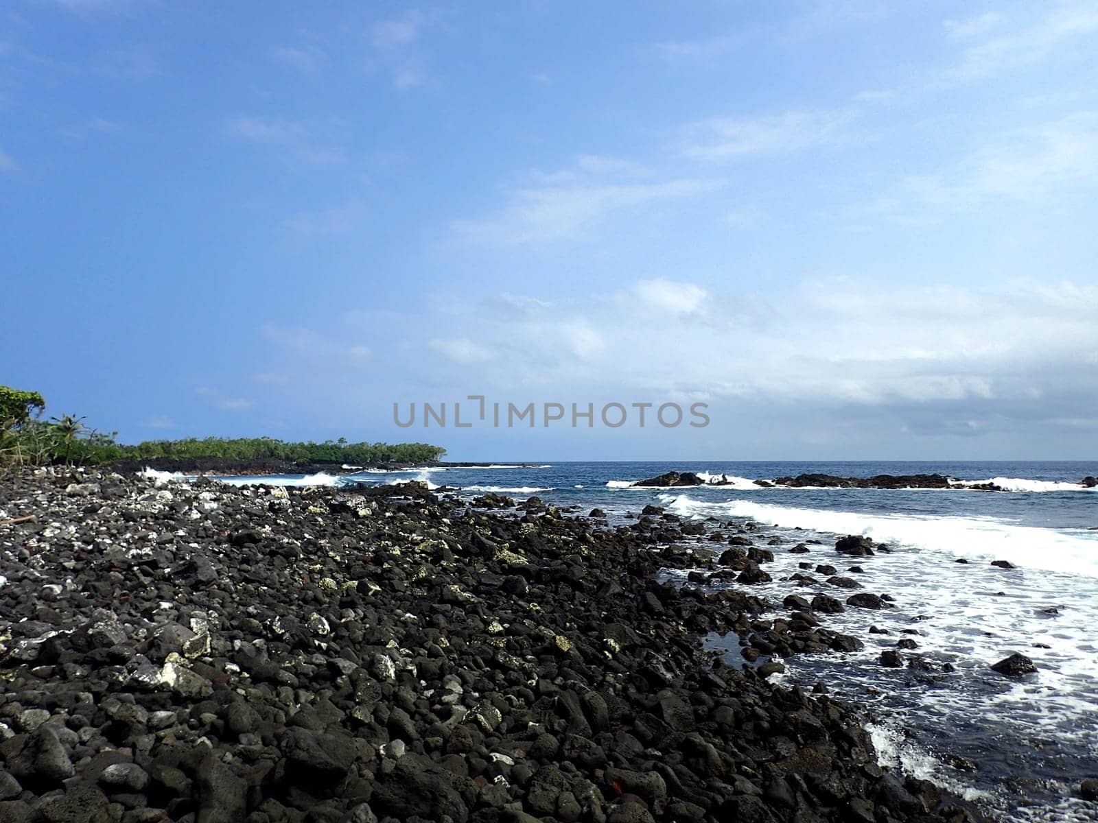 A beautiful view of the coast of Pahoa, Hawaii with blue skies and crashing waves. The photo shows the contrast between the black rocks and the white foam, and the calmness of the horizon and the movement of the water. The photo is taken from a distance, capturing the vastness and splendor of the island.