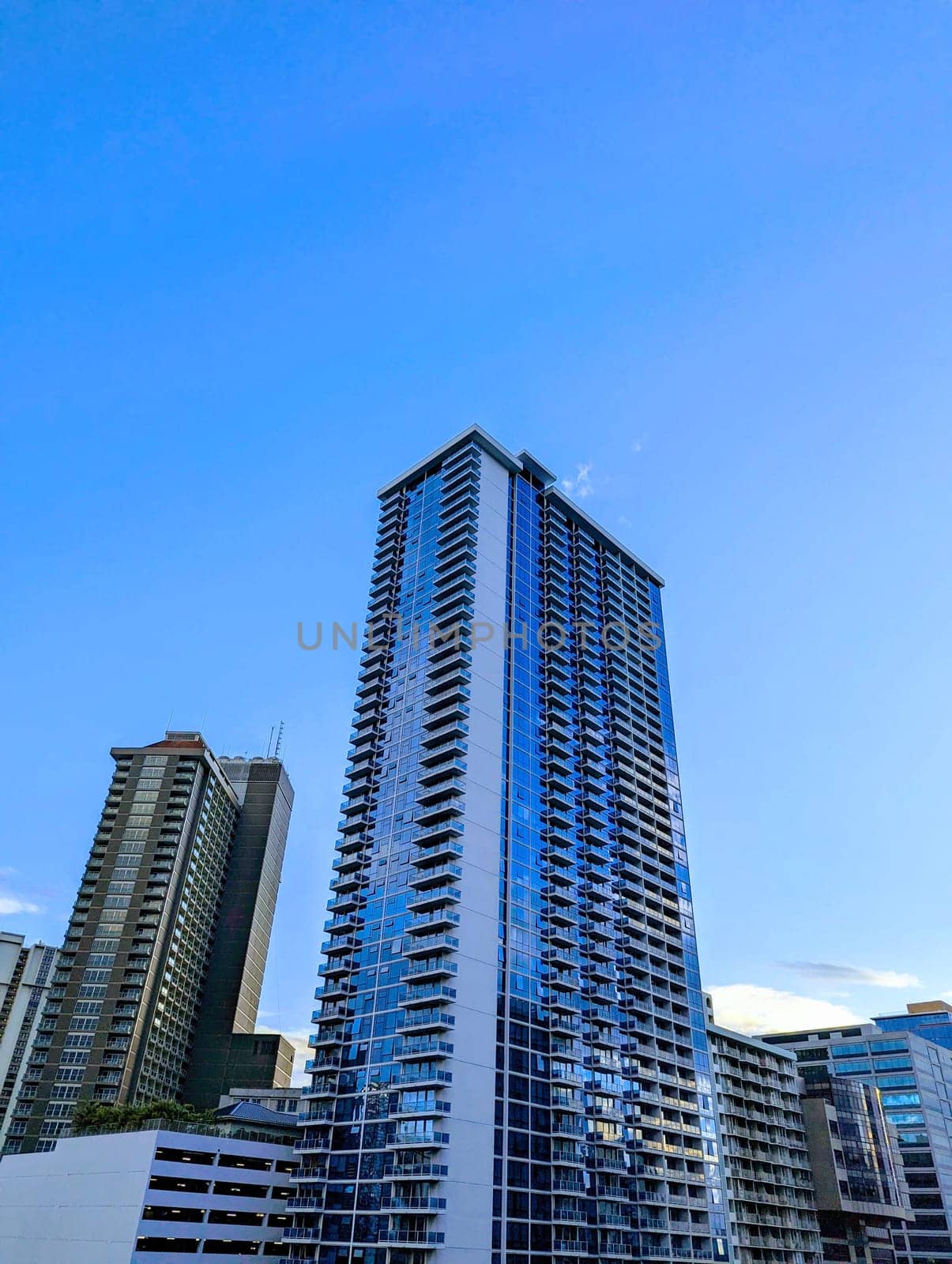 Honolulu - July 17, 2023: A photo of a modern high-rise condo building in the Ala Moana area of Oahu, Hawaii. The building is tall and rectangular with a blue glass facade. The photo shows the contrast between the urban and the natural landscape, as well as the blue sky and the clouds. The photo is taken from a low angle, looking up at the building.