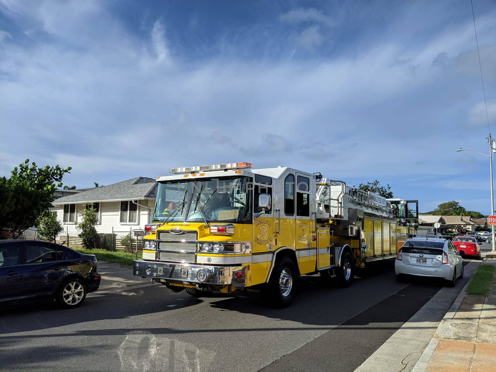 Honolulu - November 10, 2020: A fire truck driving down a small street in the Kapahulu neighborhood of Honolulu, Hawaii. The photo shows the yellow truck with a ladder on top, and the cars and houses on the sides of the street. The photo is taken from a low angle, creating a sense of movement and urgency.