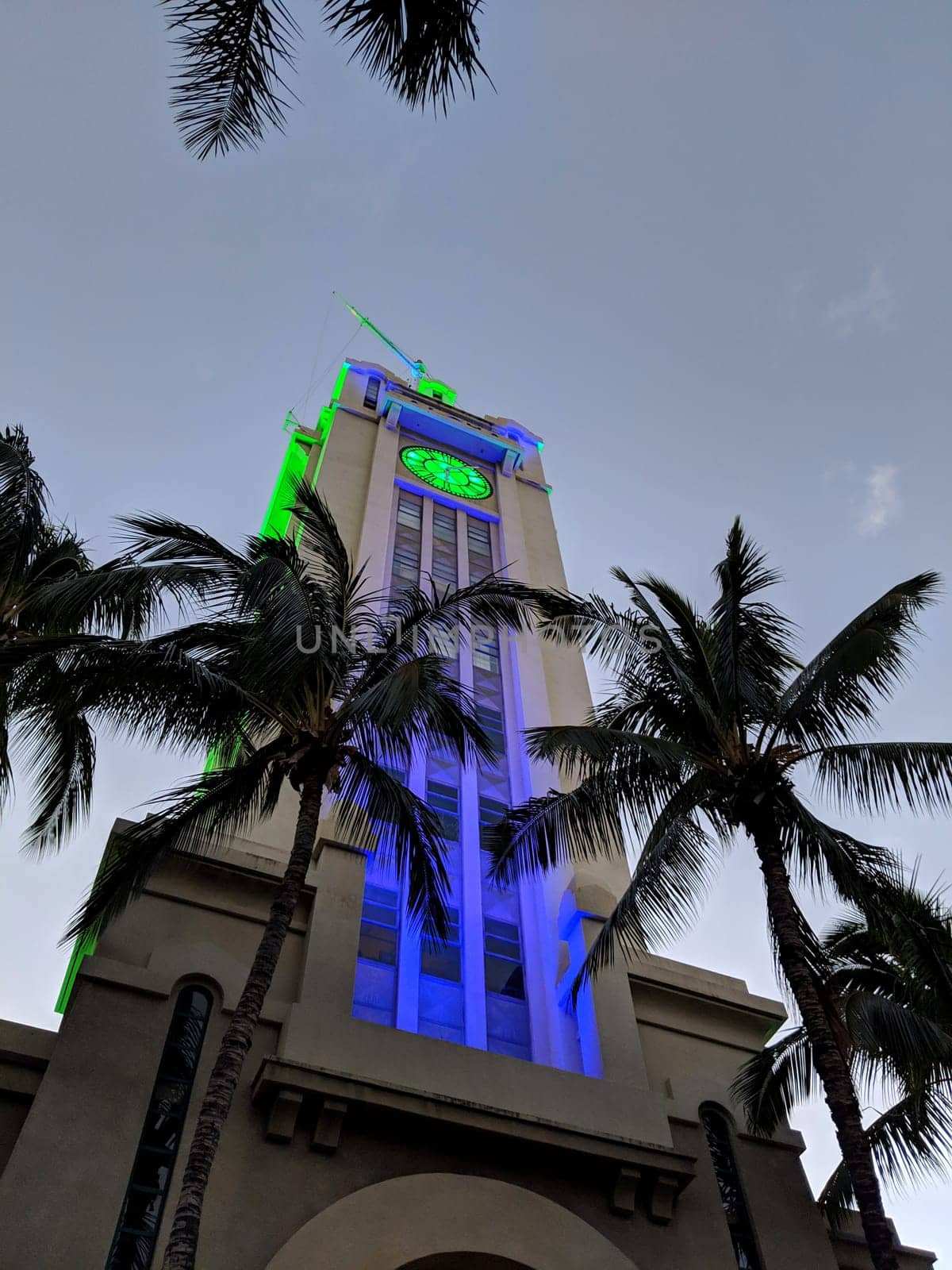 Honolulu - November 3, 2018:   Aloha Tower in Honolulu, Hawaii lit up with blue and green lights at night. The tower is framed by palm trees on either side.