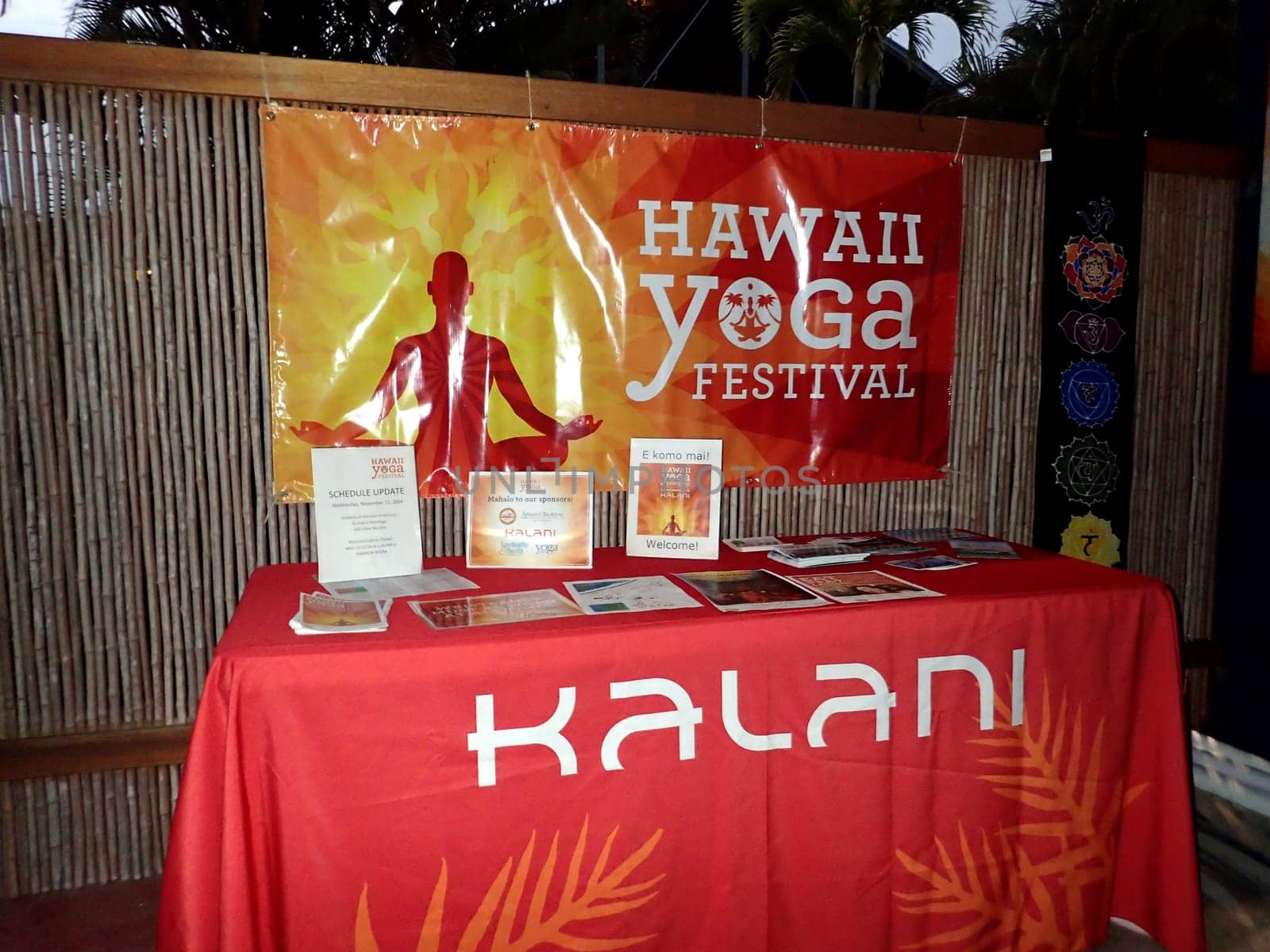 Hawaii - November 12, 2014:  banner for the Hawaii Yoga Festival, hanging on a bamboo fence at the Kalani Retreat Center in Pahoa, Hawaii. The banner is orange and yellow with a silhouette of a person in a yoga pose and the text "Hawaii Yoga Festival" in white. Below the banner is a table with a red tablecloth and various brochures and flyers, including one with the sign "Kalani" in white. The background consists of palm trees and other tropical foliage, creating a natural and serene atmosphere.