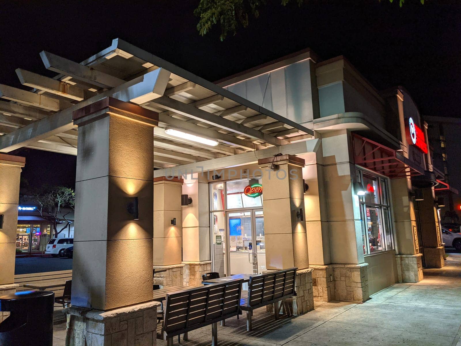 Honolulu - May 6, 2021:  Exterior of a Panda Express restaurant at night. The restaurant is a beige stucco building with a red and white sign and a neon "Open" sign. The building is lit by streetlights and the neon sign. There is a bench and a trash can in front of the building.