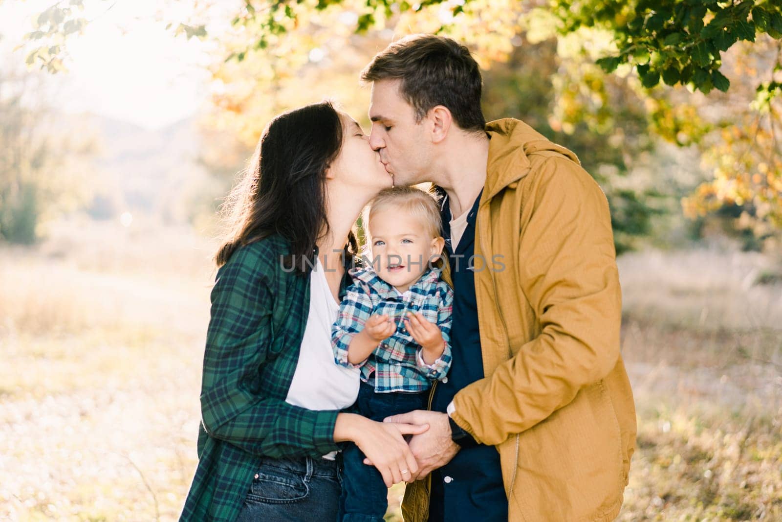 Dad kisses mom with a little girl in her arms in the park. High quality photo