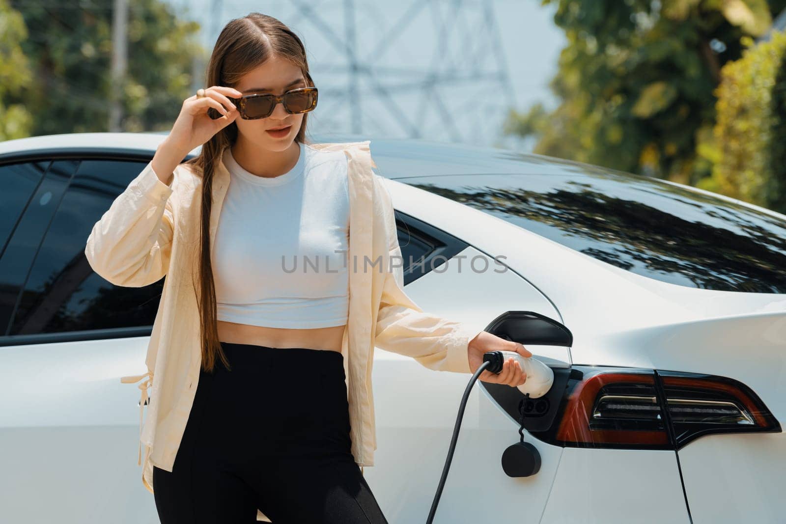 Young woman recharge her EV electric vehicle at green city park parking lot. Urban sustainability lifestyle for environmental friendly EV car with battery charging station. Expedient