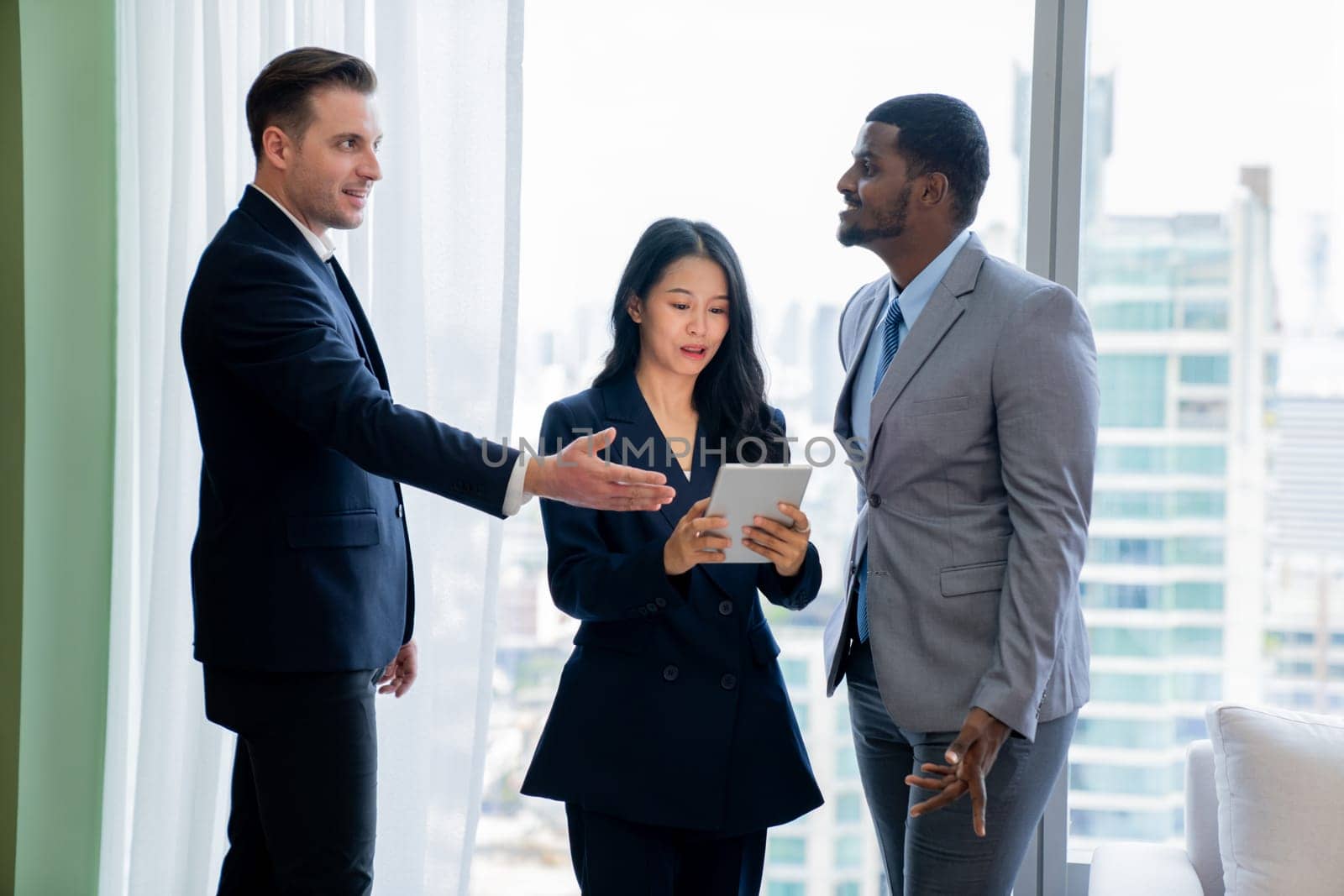 Diverse smart businesspeople standing near window with skyscraper while looking at business data analysis. Group of business team using tablet represent marketing plan, strategy, idea. Ornamented..