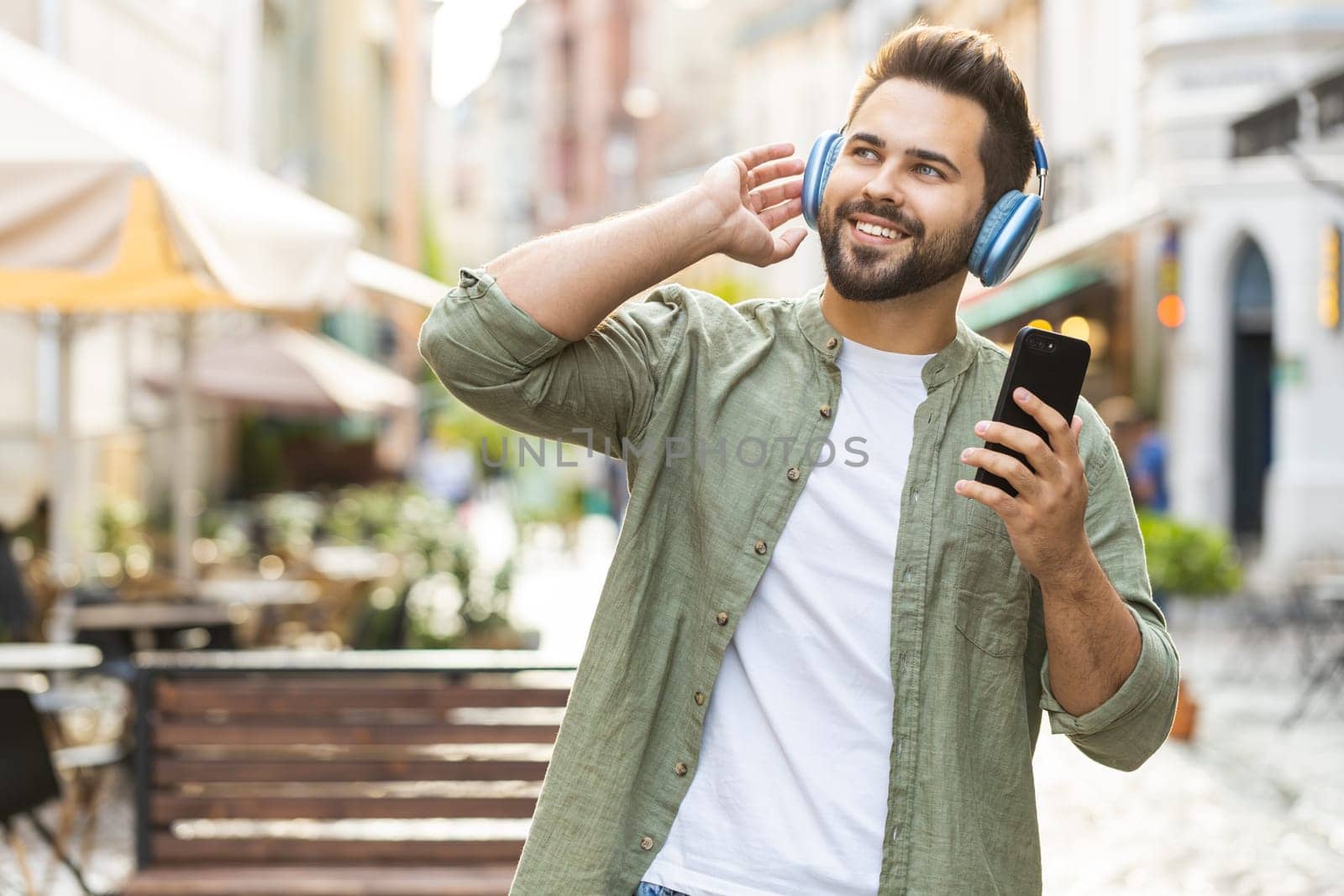 Happy relaxed overjoyed Caucasian man in wireless headphones choosing, listening favorite energetic disco music in smartphone dancing outdoors. Guy tourist walking passes by urban sunshine city street