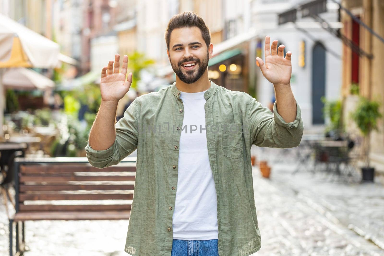 Bearded young man smiling friendly at camera, waving hands gesturing invitation hello, hi, greeting or goodbye, welcoming with hospitable expression outdoors. Guy walking in urban city town street