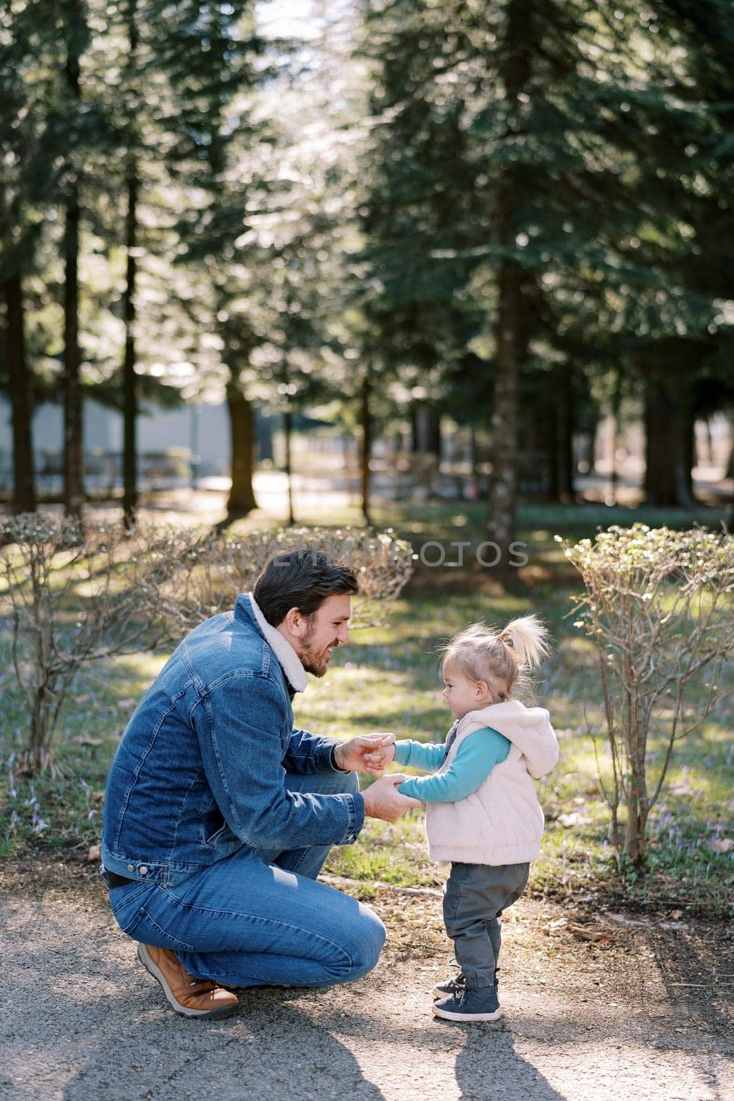 Dad holding hands a little girl squatting next to her in a spring park. High quality photo
