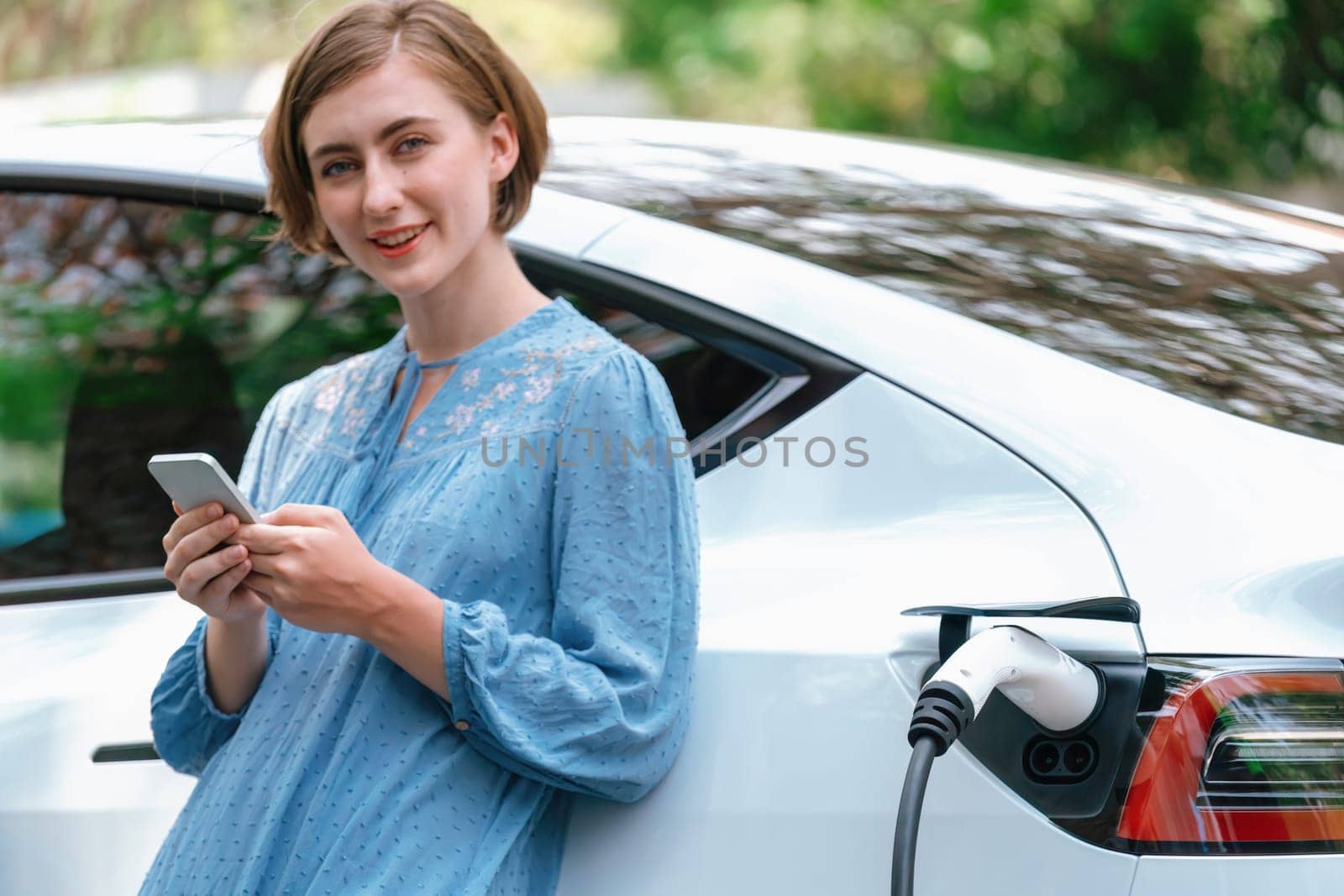 Holiday road trip vacation by the green countryside nature with beautiful young woman checking battery status from smartphone while recharging electric vehicle.Eco-friendly travel wit EV car.Perpetual