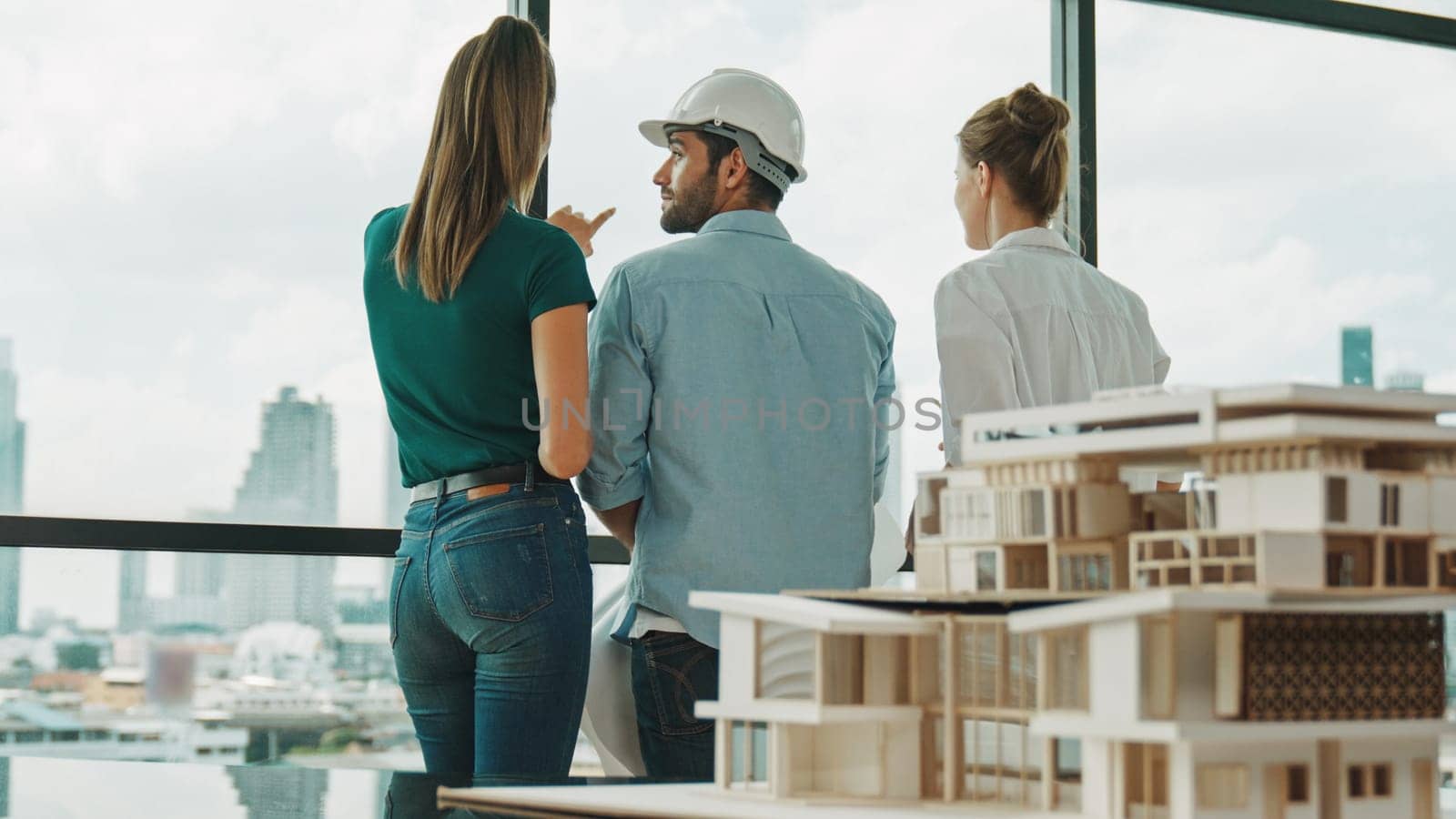 Group of caucasian diverse architect engineer pointing at skyscraper at day time. Skilled manager team looking at city or skyscraper while house model was placed on table. Back view. Tracery