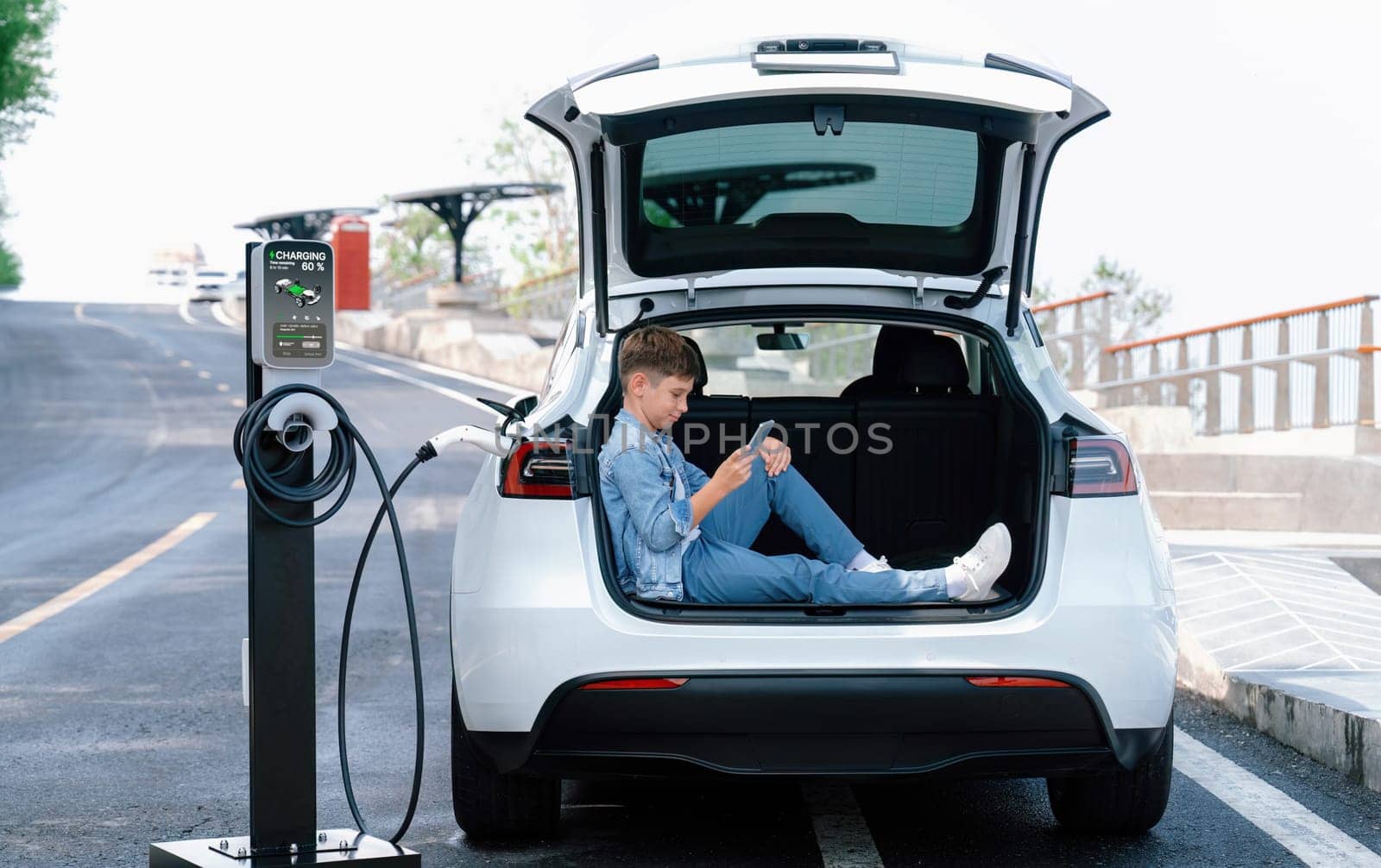 Little boy sitting on car trunk, using smartphone while recharging eco-friendly car from EV charging station. EV car road trip travel as alternative vehicle using sustainable energy concept. Perpetual