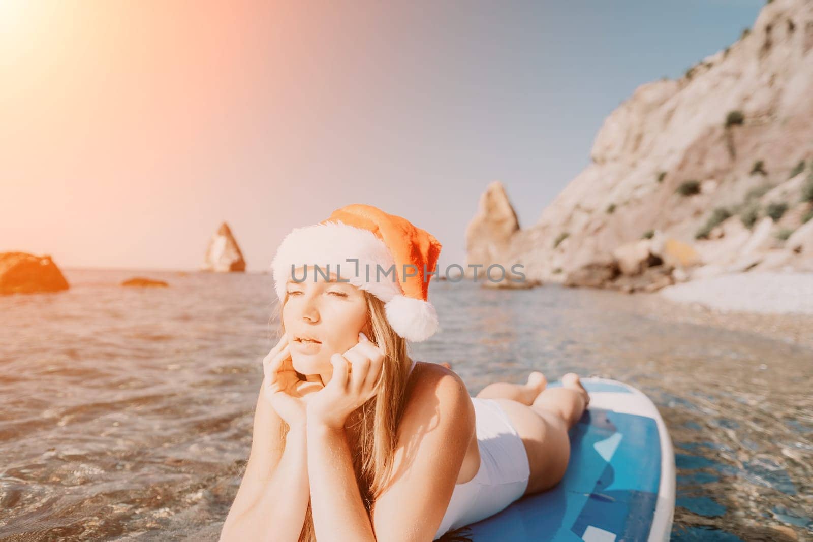 Woman sea sup. Close up portrait of happy young caucasian woman with long hair in Santa hat looking at camera and smiling. Cute woman portrait in a white bikini posing on sup board in the sea by panophotograph