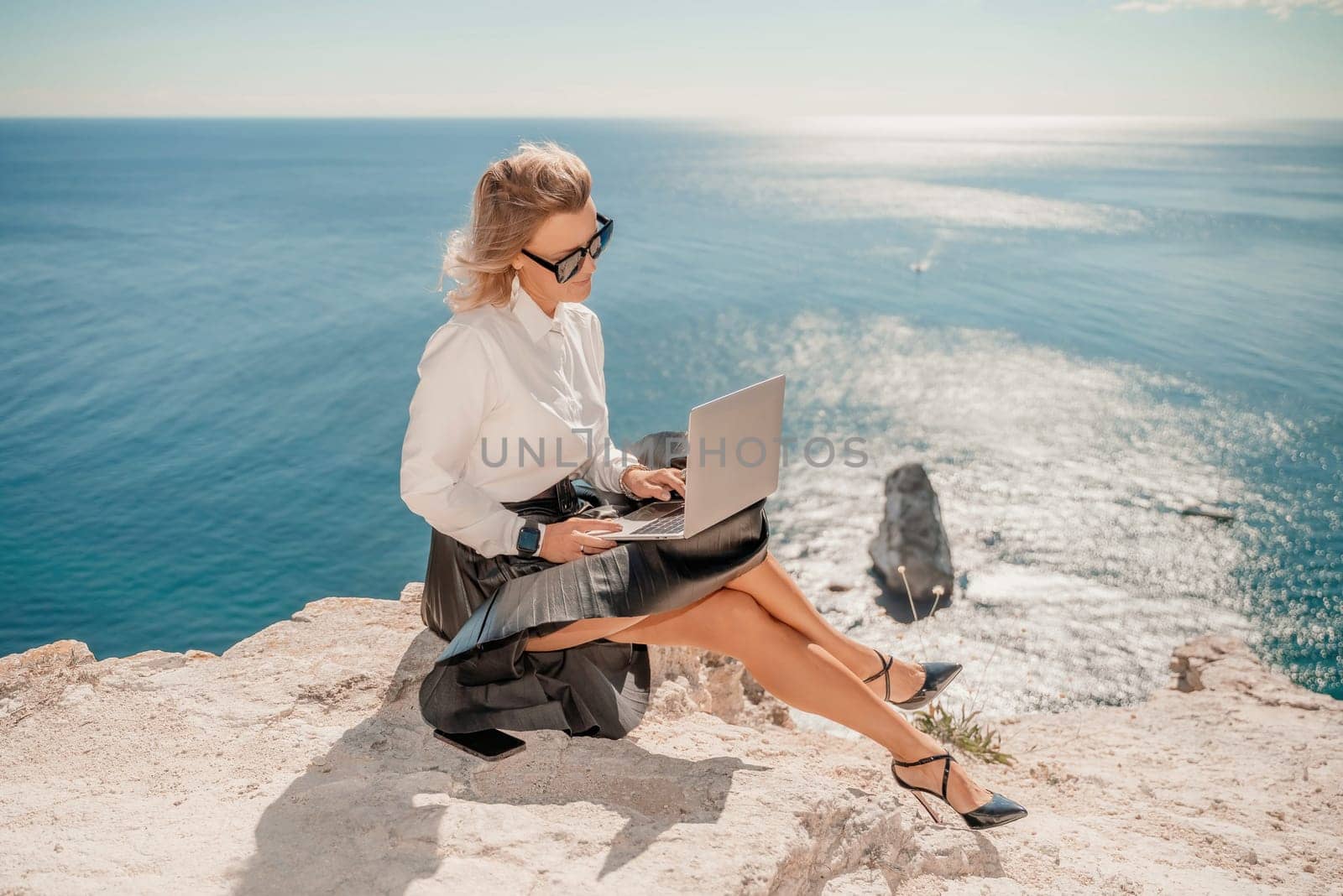 Business woman on nature in white shirt and black skirt. She works with an iPad in the open air with a beautiful view of the sea. The concept of remote work