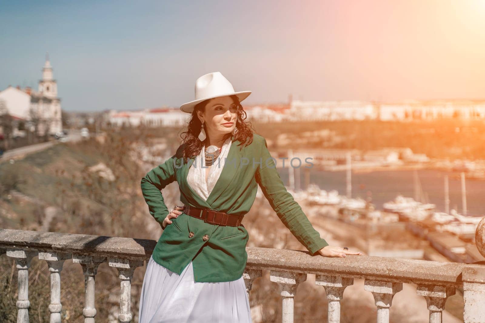 Woman walks around the city, lifestyle. Happy woman in a green jacket, white skirt and hat is sitting on a white fence with balusters overlooking the sea bay and the city. by Matiunina