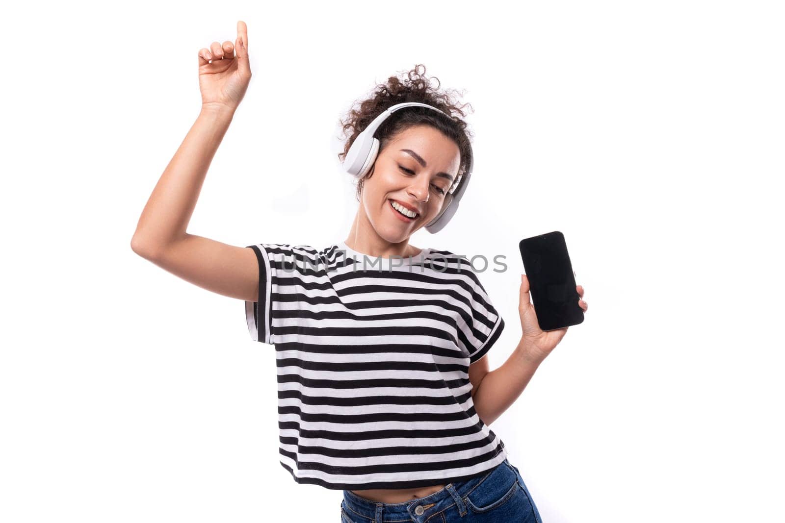 young caucasian woman with curly haircut dressed in a striped summer t-shirt with wireless headphones shows ads on a smartphone screen with a mockup.