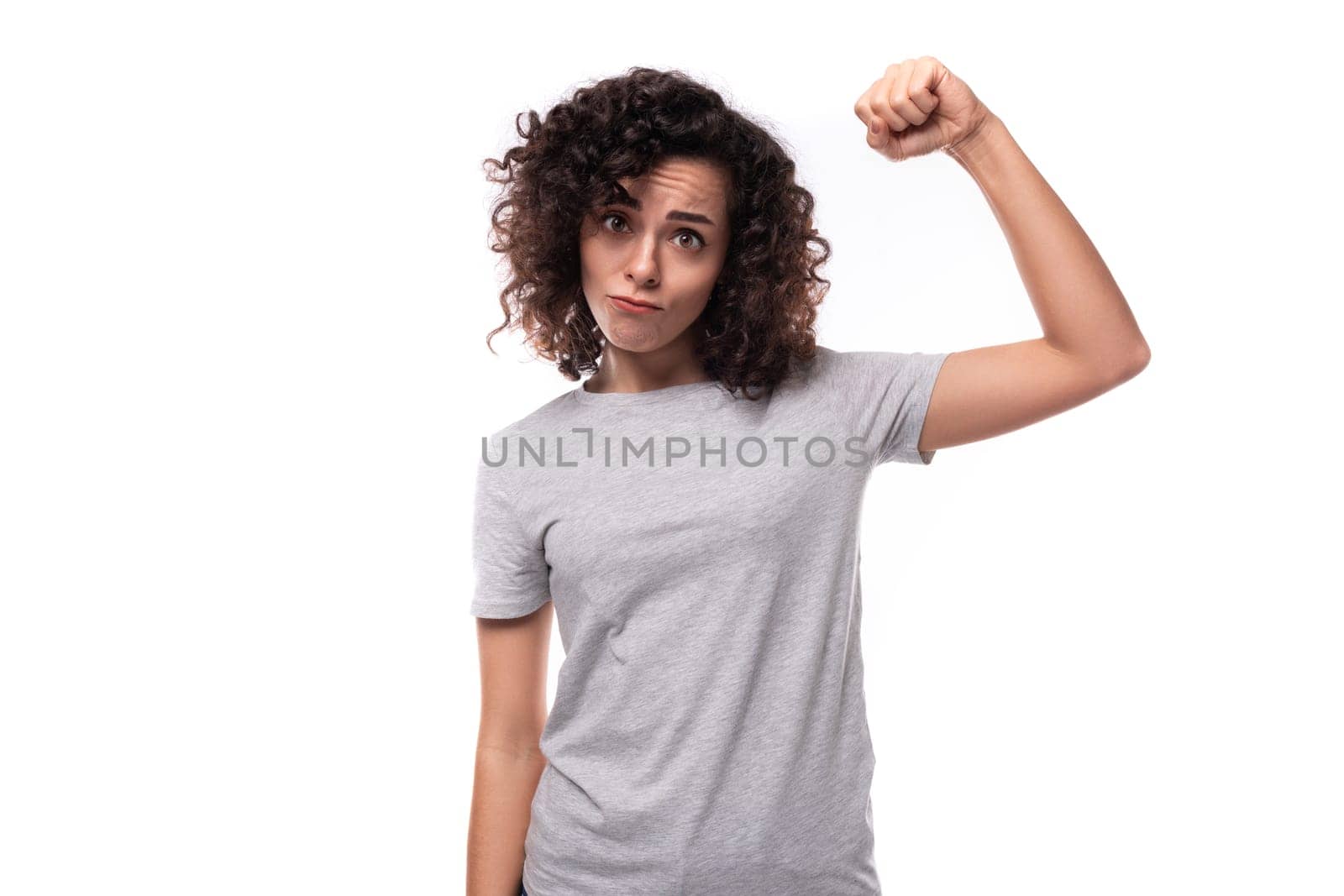 pretty confident positive young caucasian woman with black curly hair is dressed in a gray t-shirt on a white background with copy space by TRMK