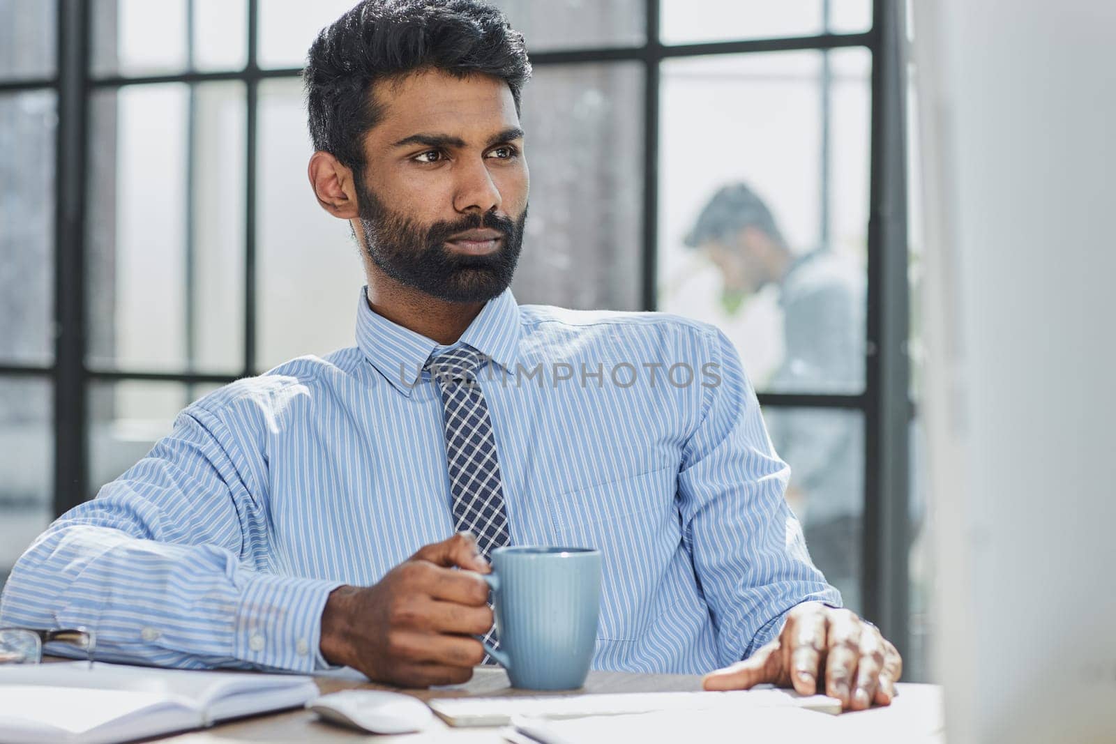 A handsome young man is sitting by the window in the office, drinking coffee while working on a laptop.