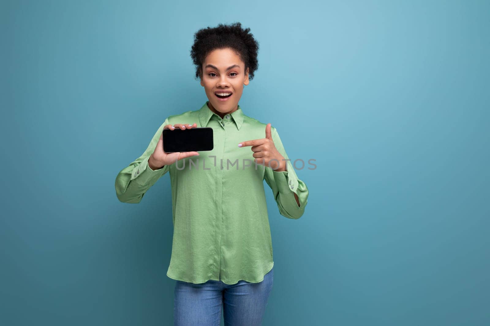young surprised hispanic brunette lady dressed in a green stylish shirt demonstrates the smartphone screen on the background with copy space.
