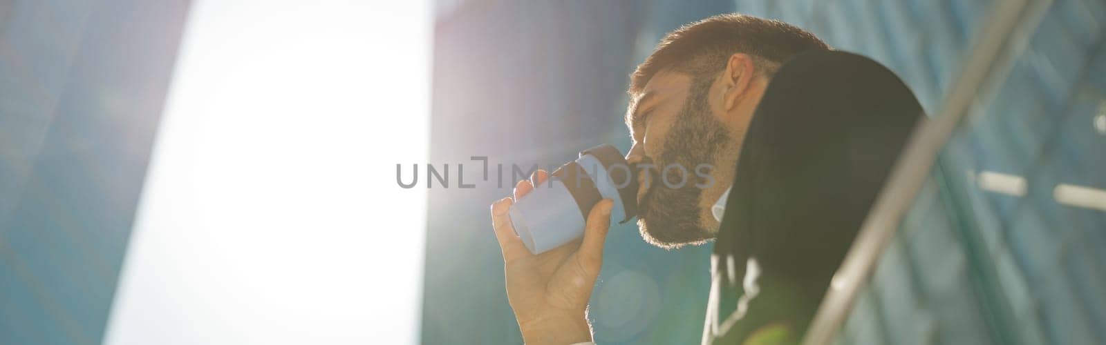 Handsome businessman with phone is drinking coffee standing on background of city skyscrapers