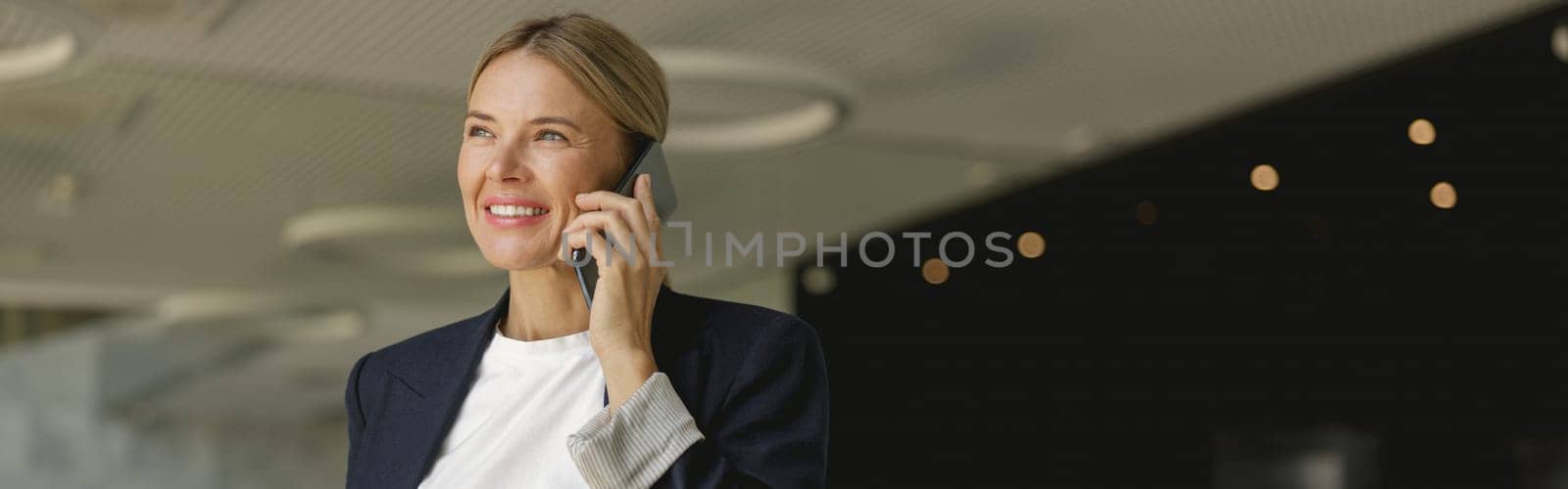 Cheerful businesswoman talking phone sitting on modern office background and looks away