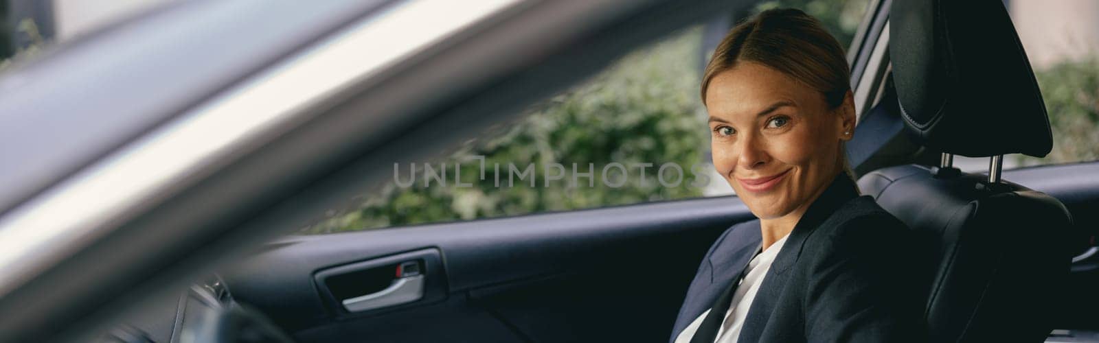 Smiling female manager is fastening car seat belt while sitting on passenger seat and looks camera