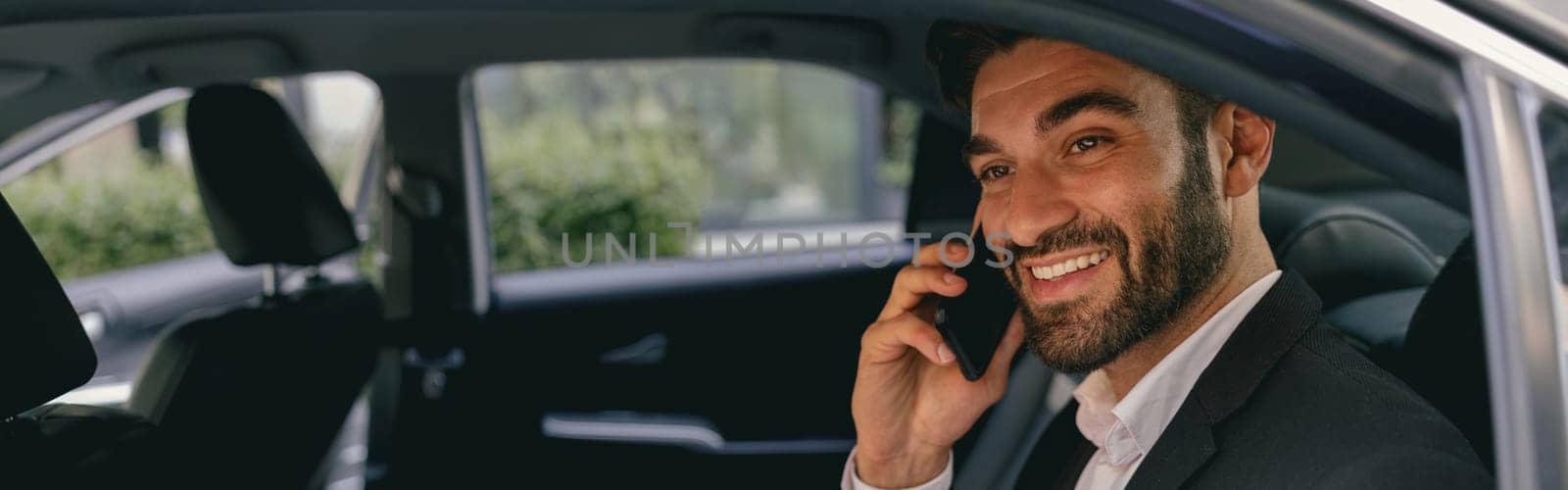 Handsome smiling businessman in suit using personal computer and making phone call in taxi by Yaroslav_astakhov