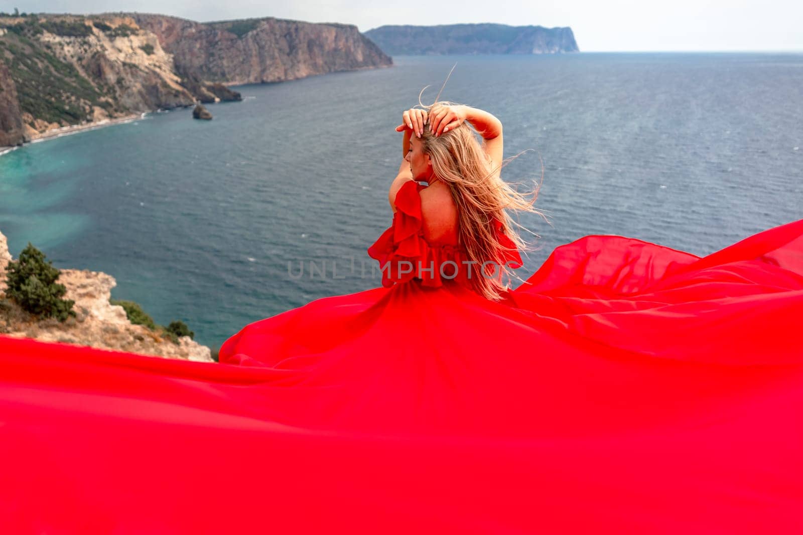 A woman in a red silk dress sits by the ocean with mountains in the background, her dress swaying in the wind. by Matiunina
