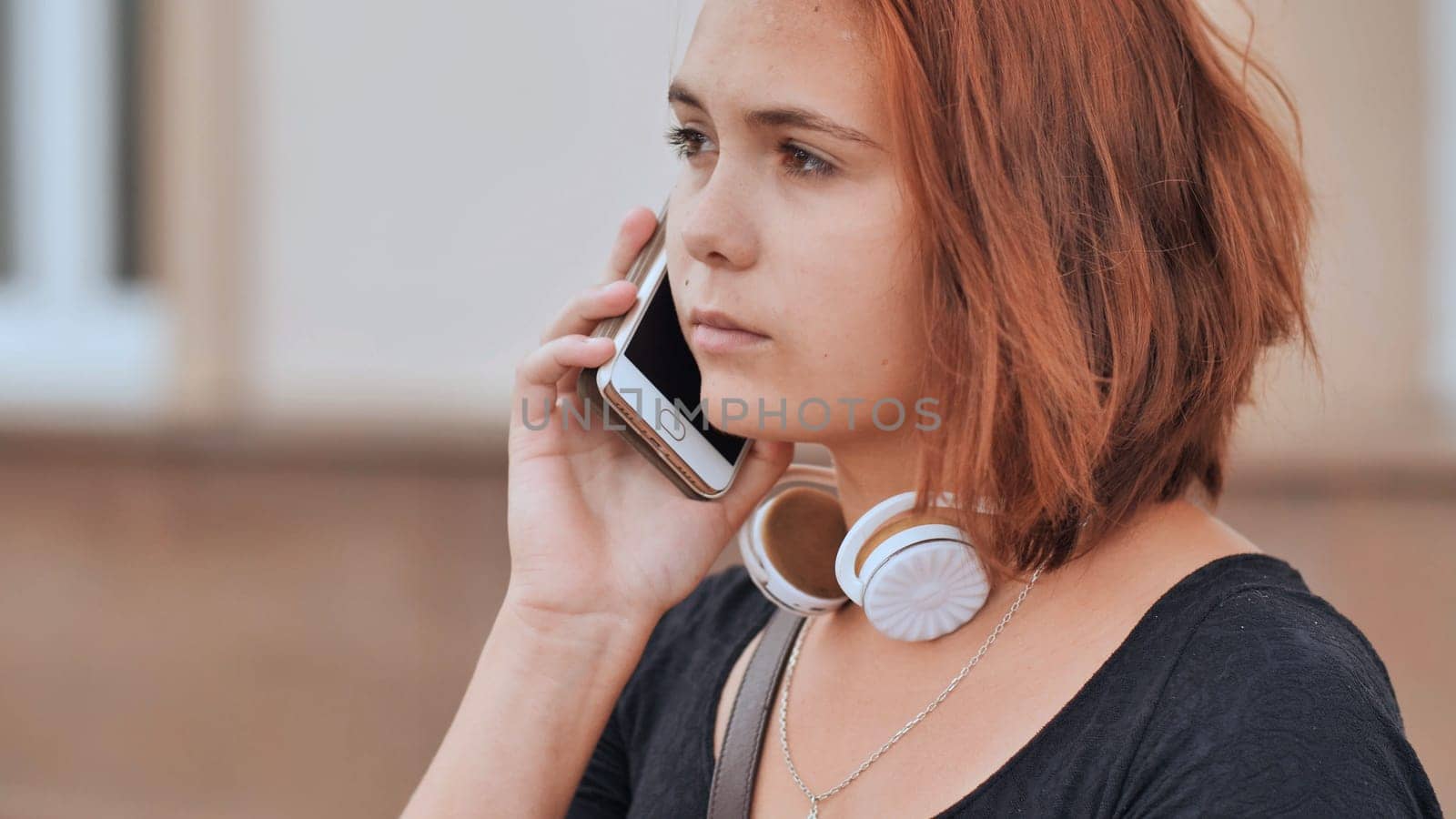 Sad red-haired girl in headphones dials a number on the phone and waits for an answer