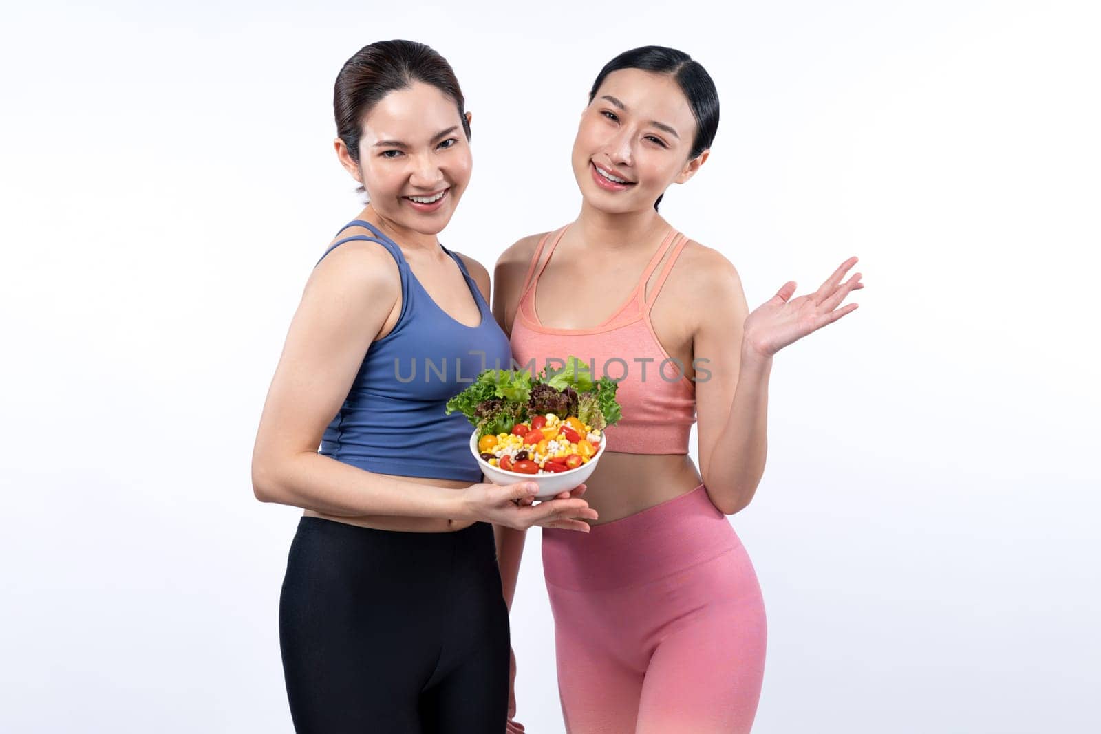 Two young sporty Asian women in sportswear holding salad bowl fill with fruit and vegetable. Natural youthful and fit body lifestyle people with balance nutrition on isolated background. Vigorous