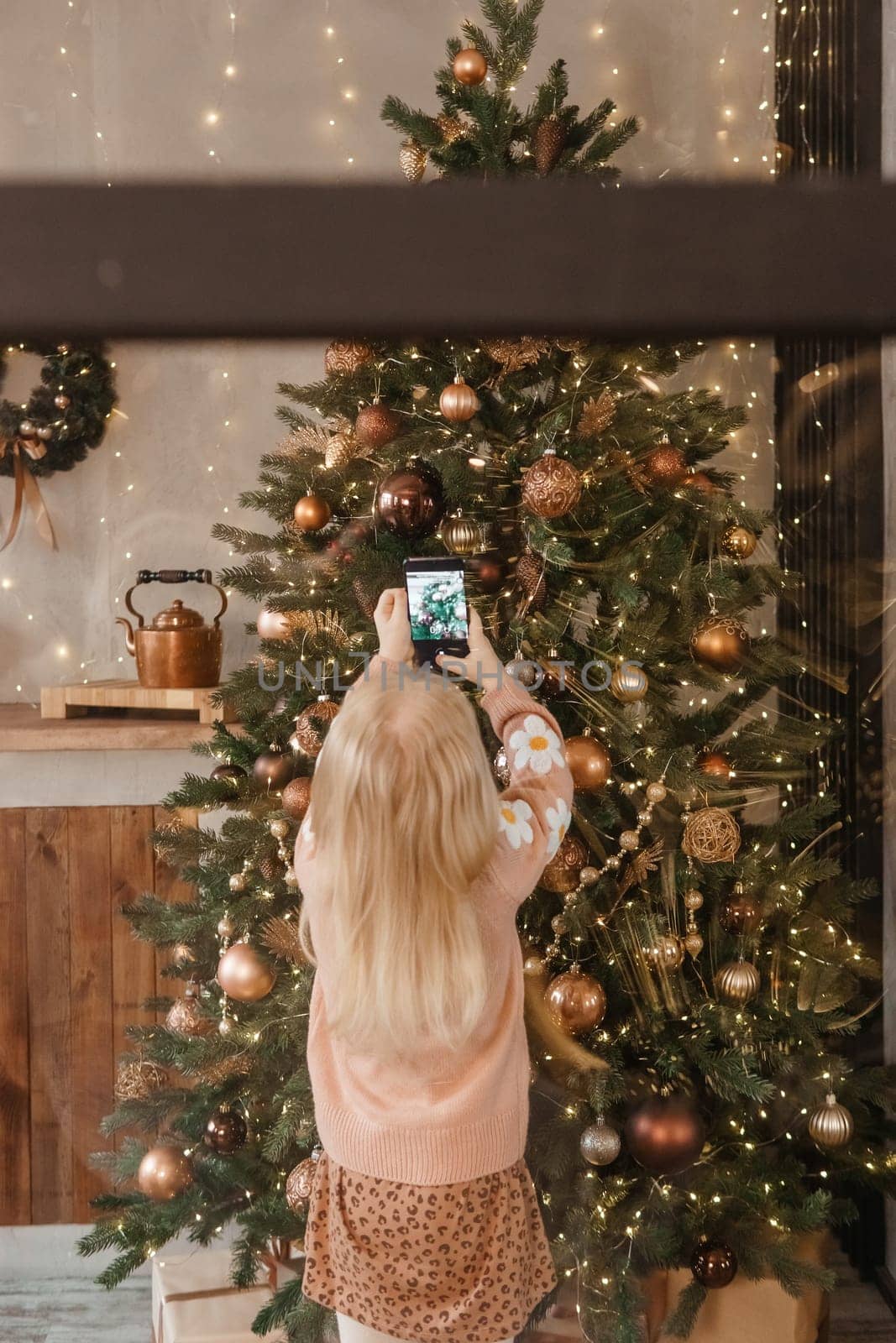 A little blonde girl photographs balloons on a Christmas tree in a festive interior decorated in a New Year's style. The concept of a merry Christmas