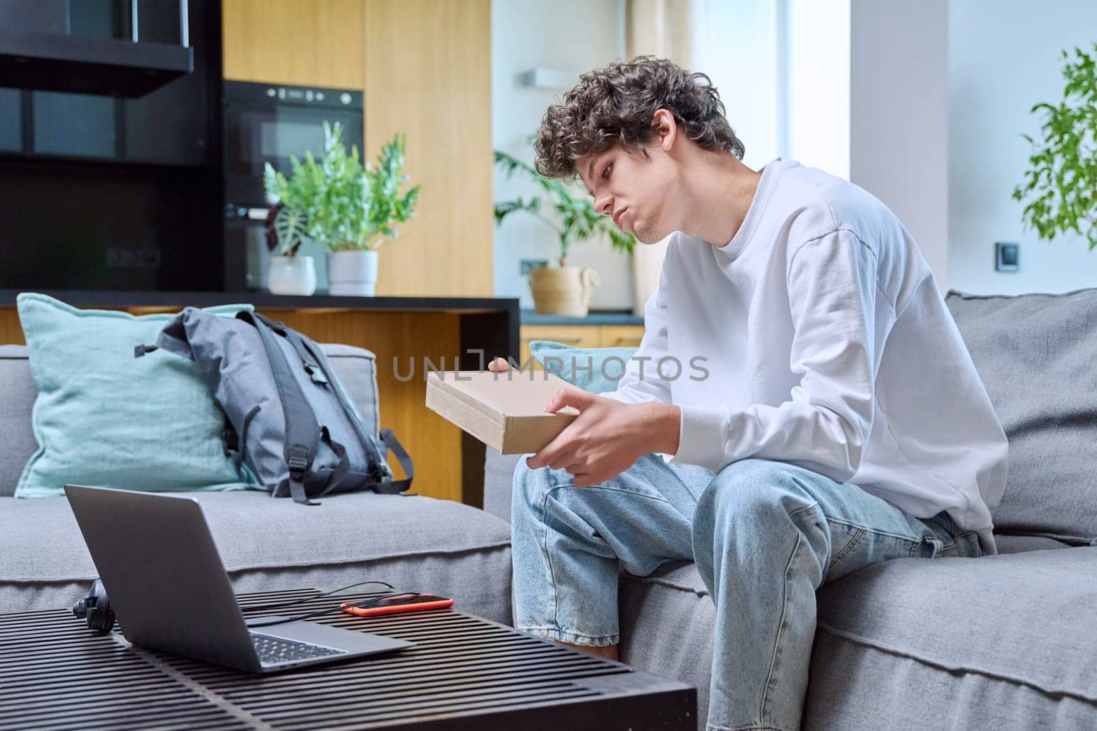 Young male unpacking an online purchase, sitting on couch at home by VH-studio