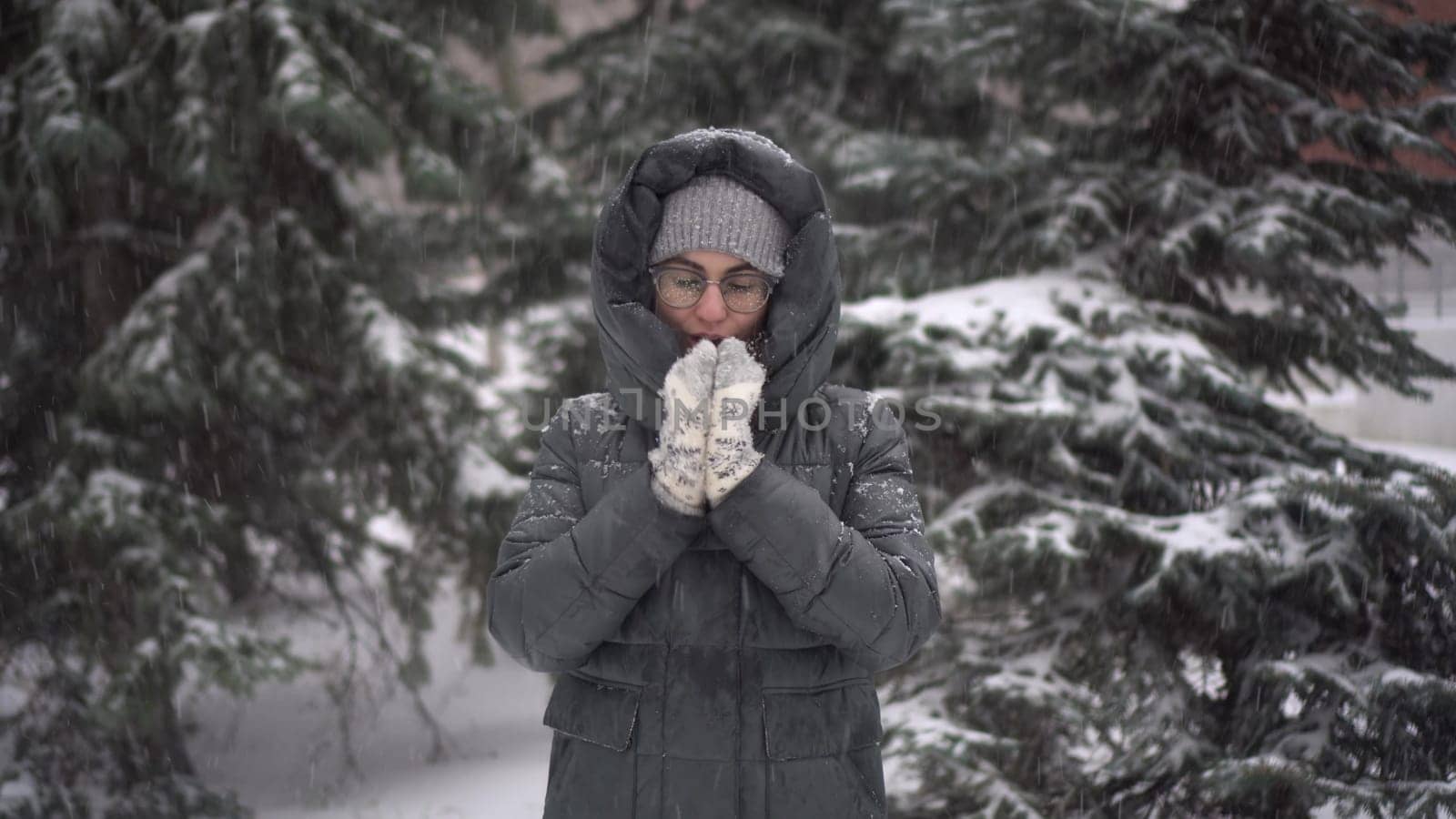 A young woman stands against a background of fir trees under heavy snowfall and rubs her hands against the cold. A girl in glasses and a down jacket with a hood stands and looks at the camera. by Puzankov