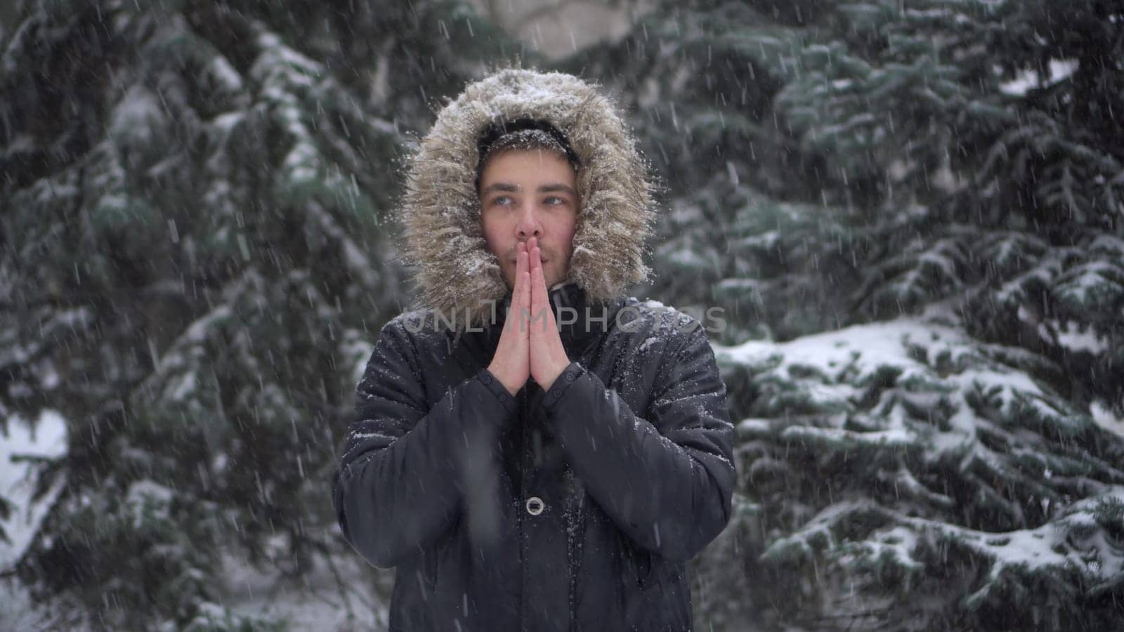 A young man stands against a background of fir trees under heavy snowfall and rubs his hands against the cold. A man in a down jacket with a hood stands and looks at the camera. 4k
