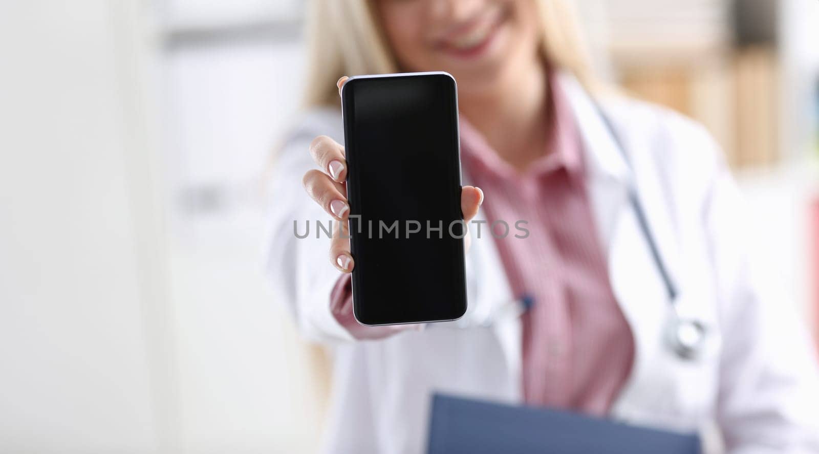 Beautiful female blonde doctor holding smartphone in hand at office woman discussing disease and giving an online consultation to a remote training mobile advertising app