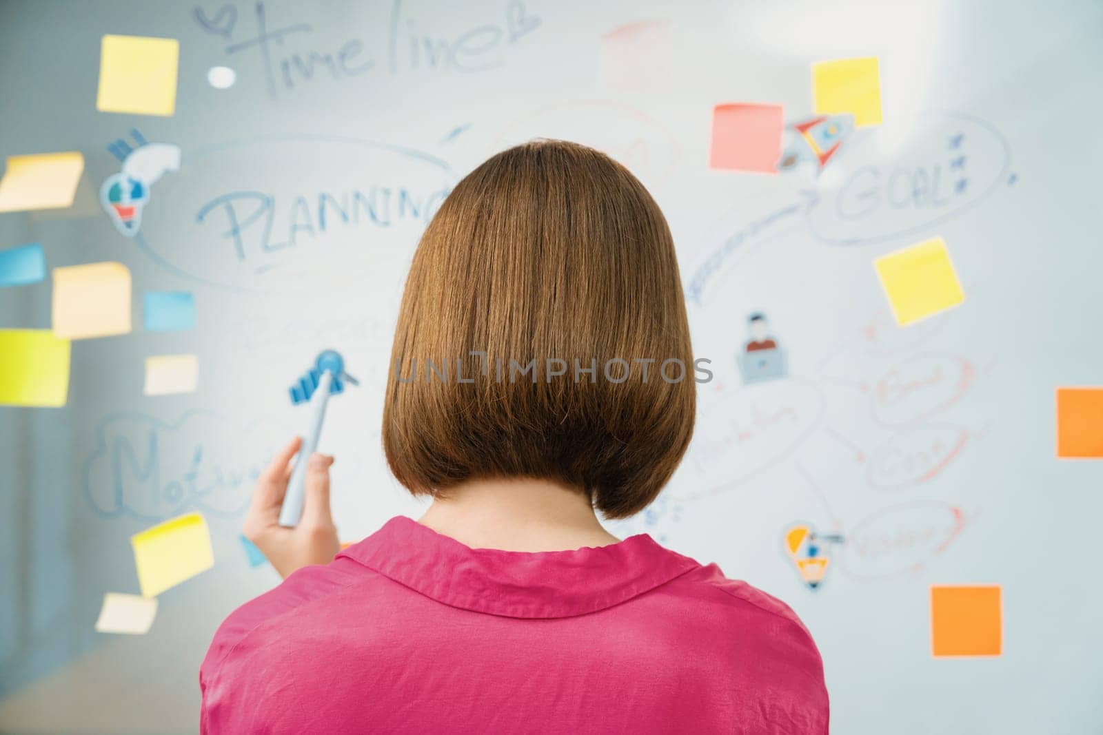 Beautiful businesswoman putting sticker on glass board while thinking about creative marketing strategy by using mind map and sticky note. Creative business meeting concept. Back view. Immaculate.