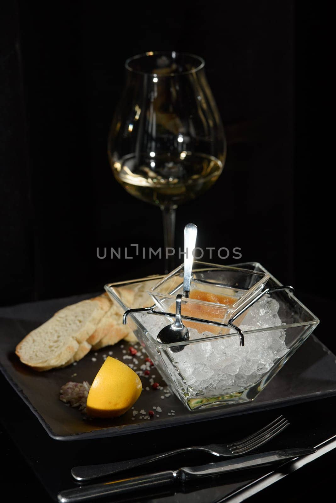 Pike fish caviar, on ice, with croutons and butter, on a transparent dish with a glass of white wine on a dark background