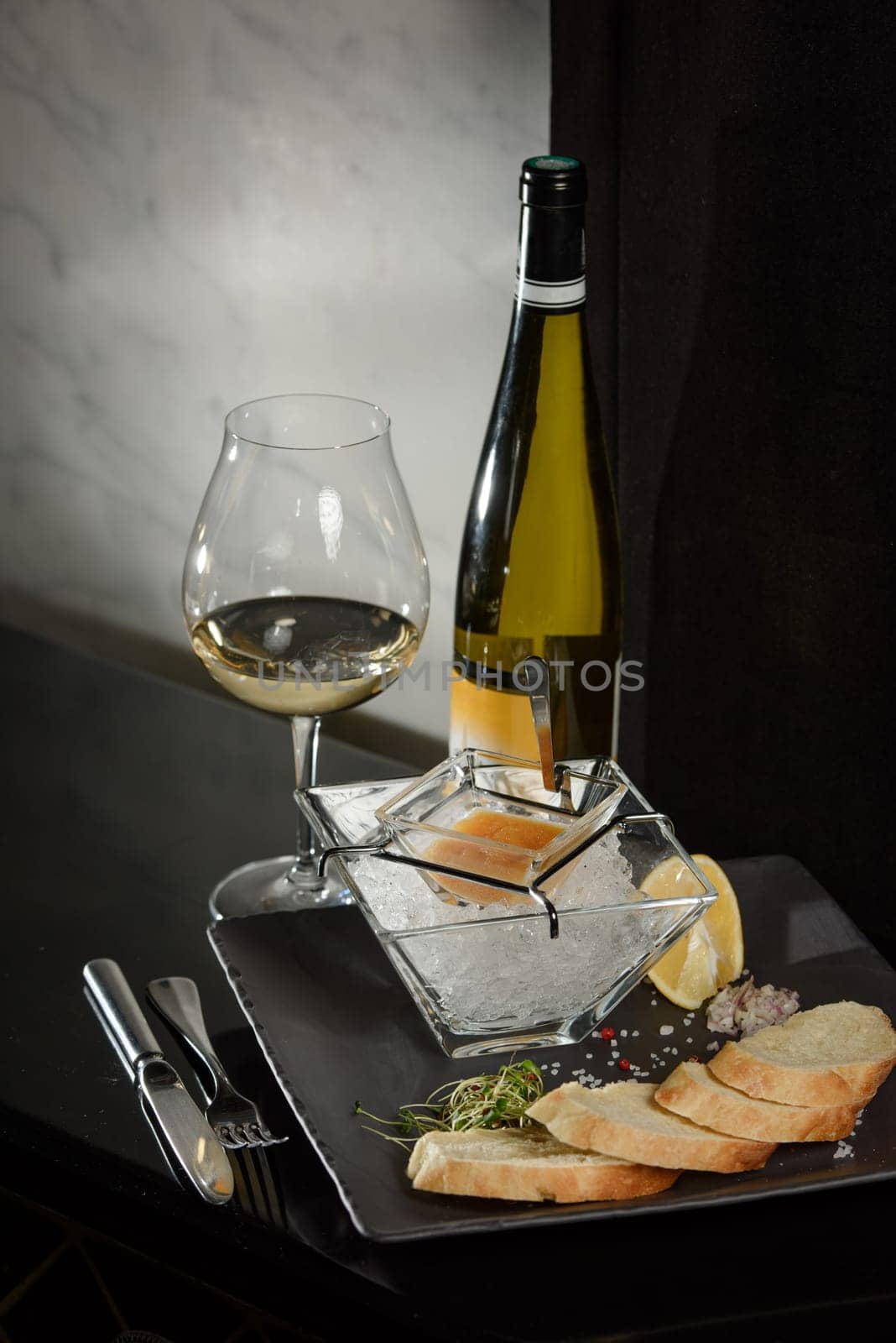 Pike fish caviar, on ice, with croutons and butter, on a transparent dish with a glass of white wine on a dark background