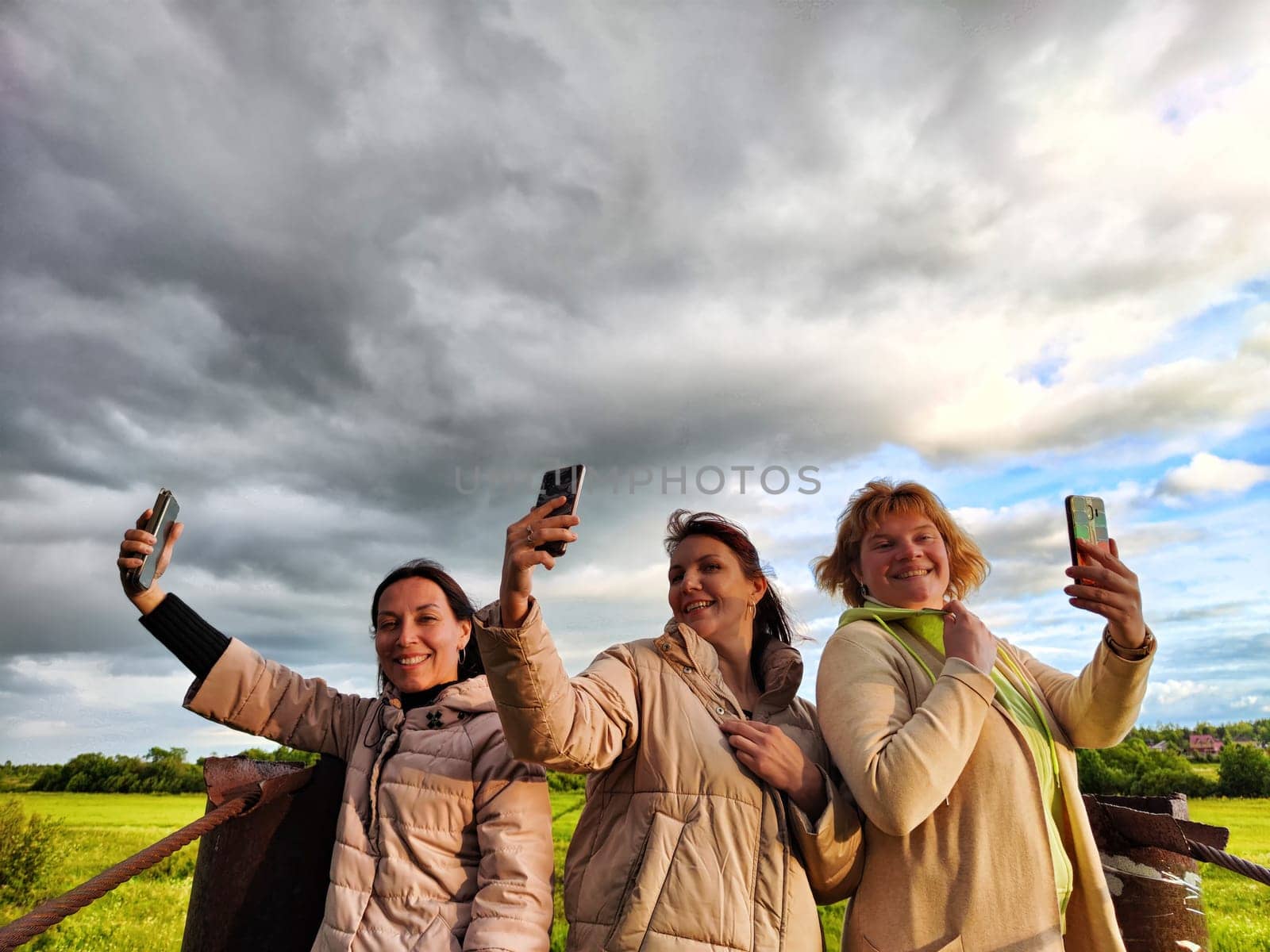 Three funny tourist girls on the old bridge take selfies against the background of an alarming dramatic sky with thunderclouds. Middle-aged women having fun outdoors by keleny