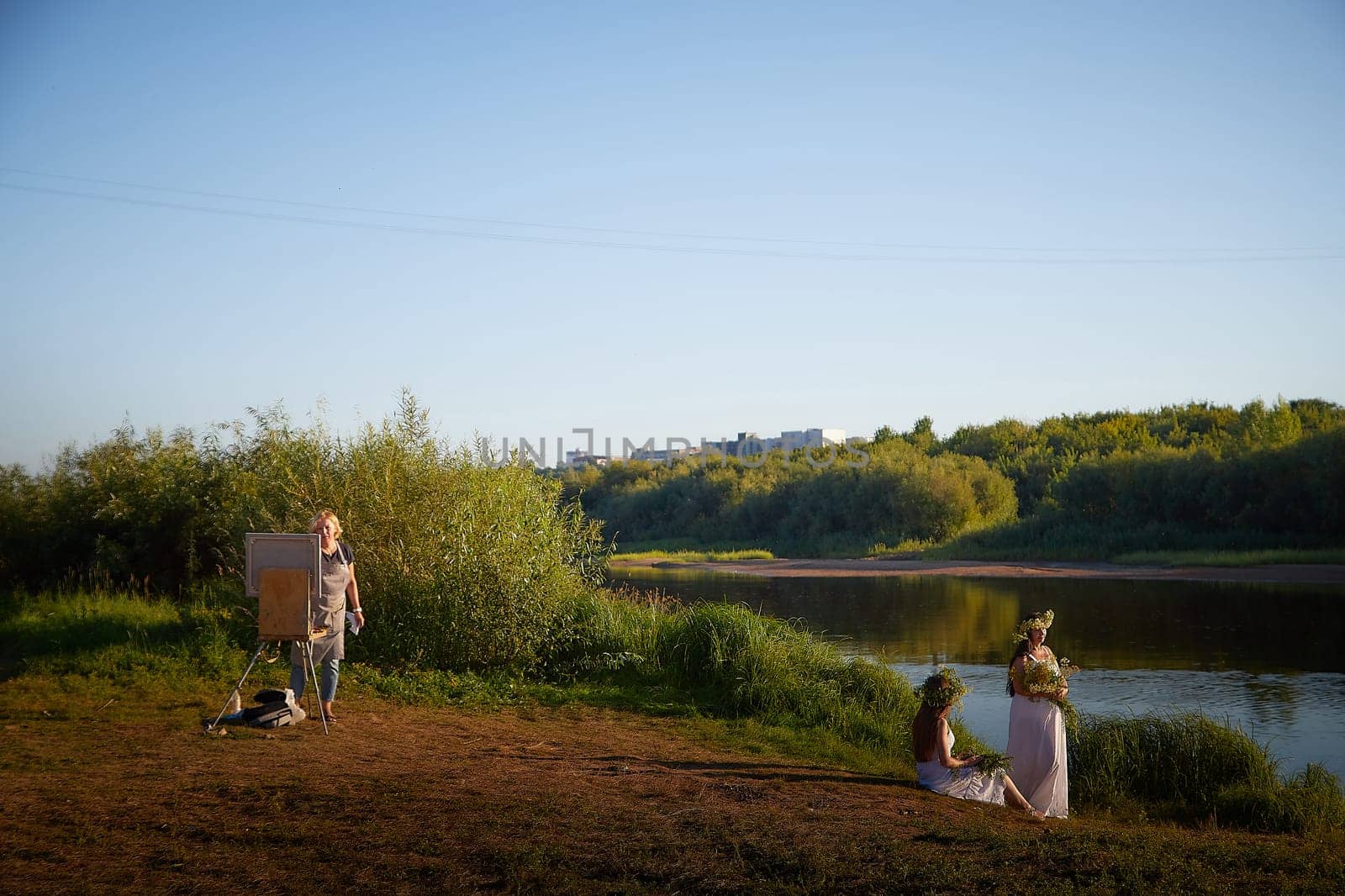 Adult female artist painting picture near water of river or lake in nature and girls in white sundress and flower wreath posing in the holiday of Ivan Kupala in nature at sunset by keleny