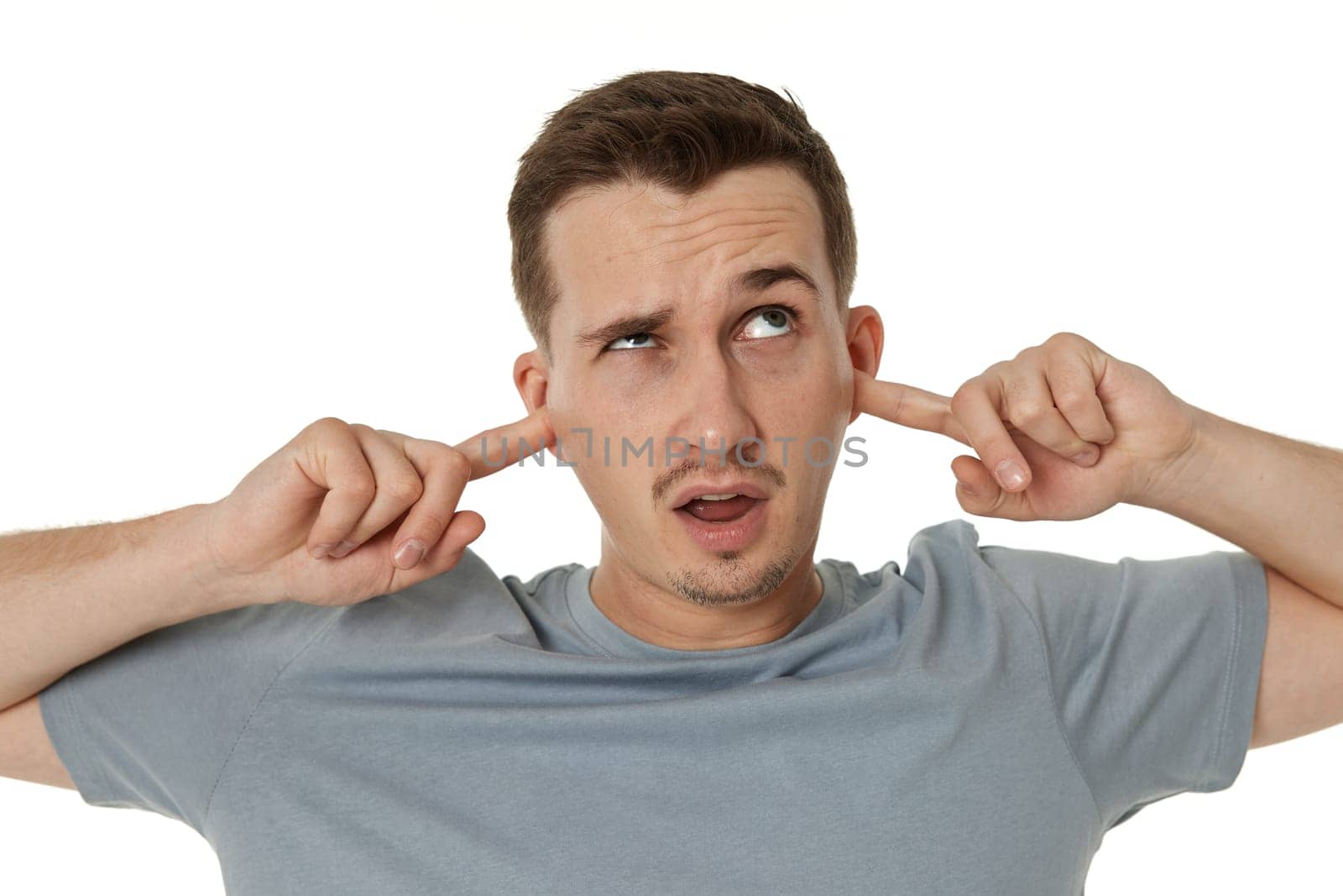 Young annoyed caucasian man in t-shirt covers ears with hands fingers on white background