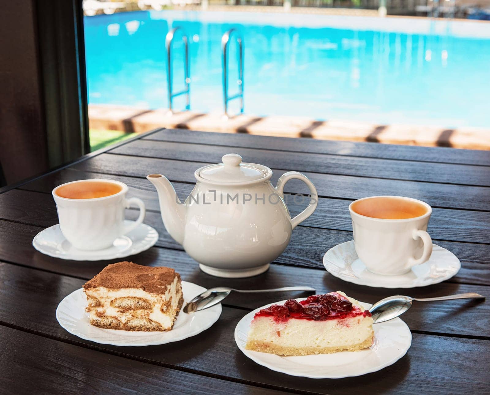 Afternoon tea set with hot tea and tasty cakes on the table and the swimming pool. Hotel resort or resting concept